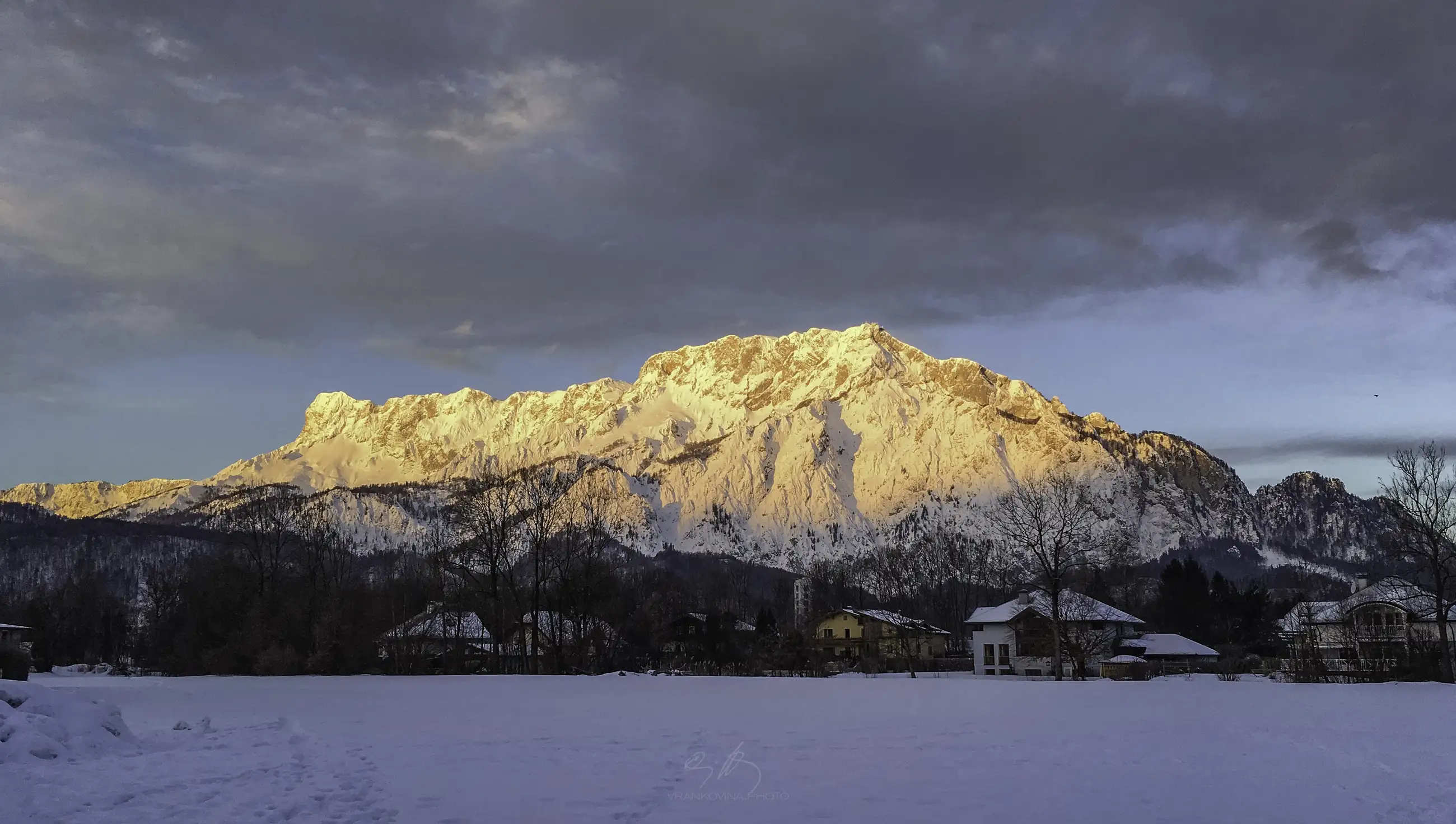 Snow-covered mountains bathed in golden sunlight under a partly cloudy sky, with a snowy field and scattered houses in the foreground.