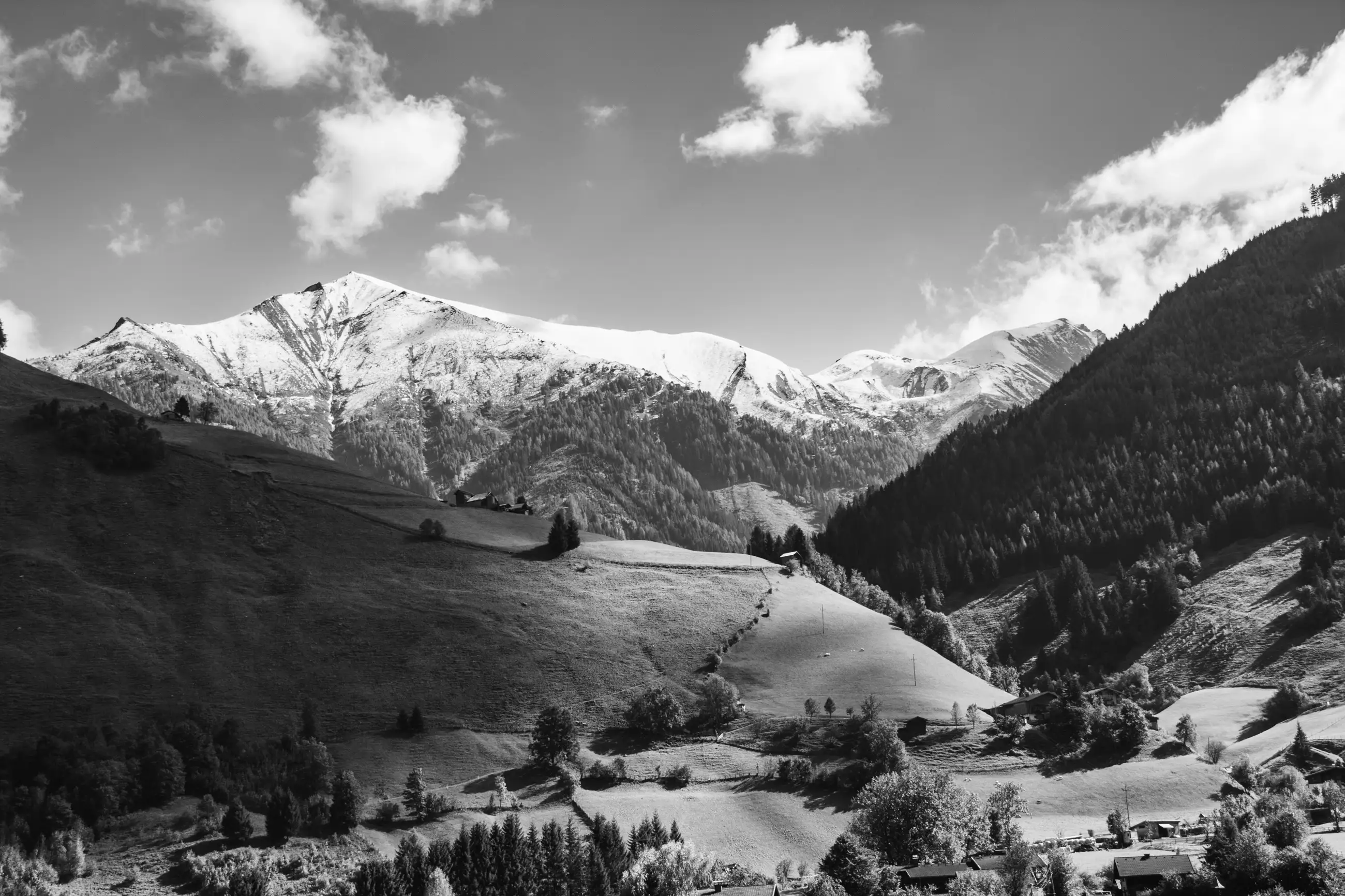 Black and white image of a scenic mountain landscape with snow-capped peaks, rolling green hills, scattered trees, and a few buildings nestled at the base of the hills under a partly cloudy sky.