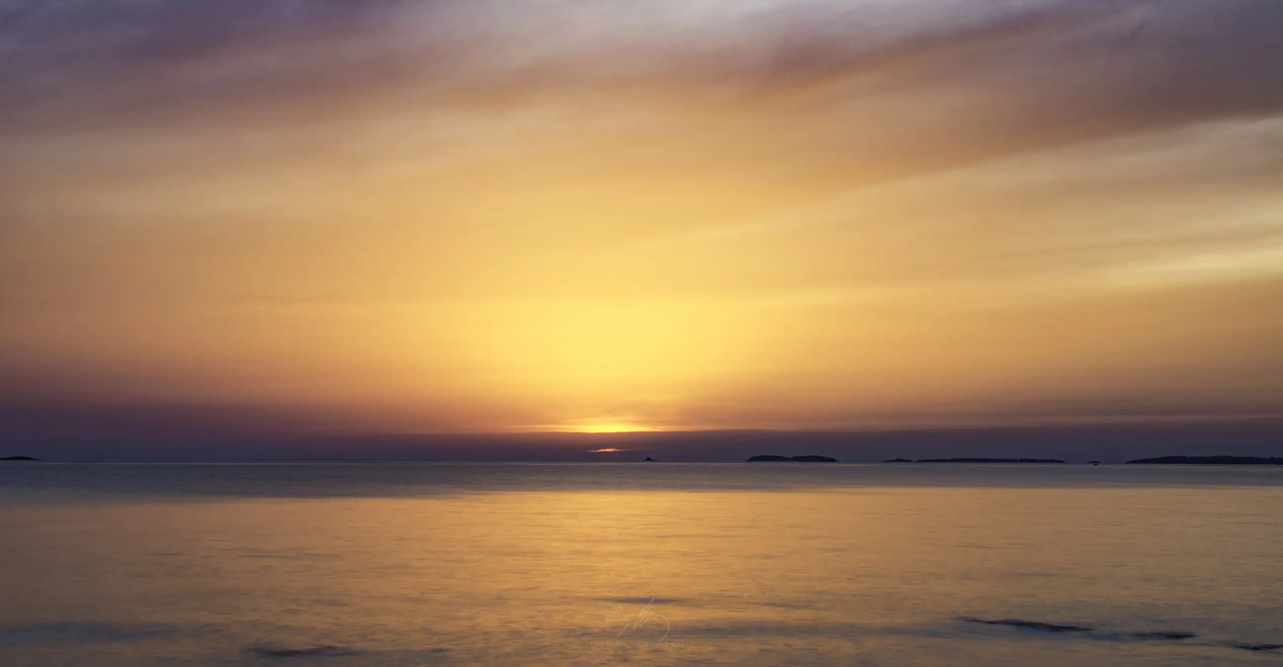 Calm sea at sunset with the sun low on the horizon, casting orange, yellow, and pink hues across the sky and water. A few small, dark islands are visible in the distance.