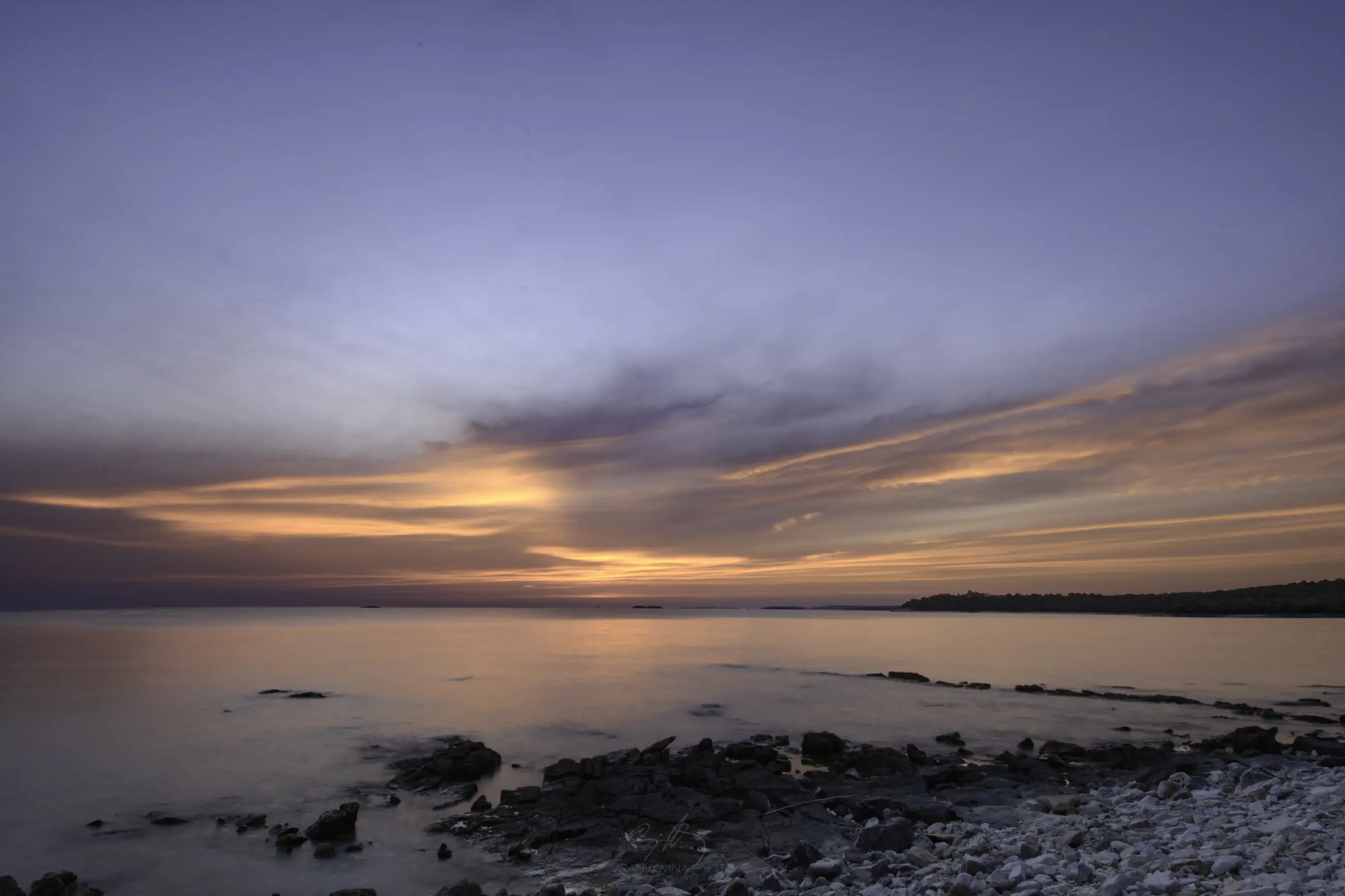 A rocky shoreline at sunset with calm water reflecting the colorful sky, which features shades of orange, pink, purple, and blue, blending together above the horizon.