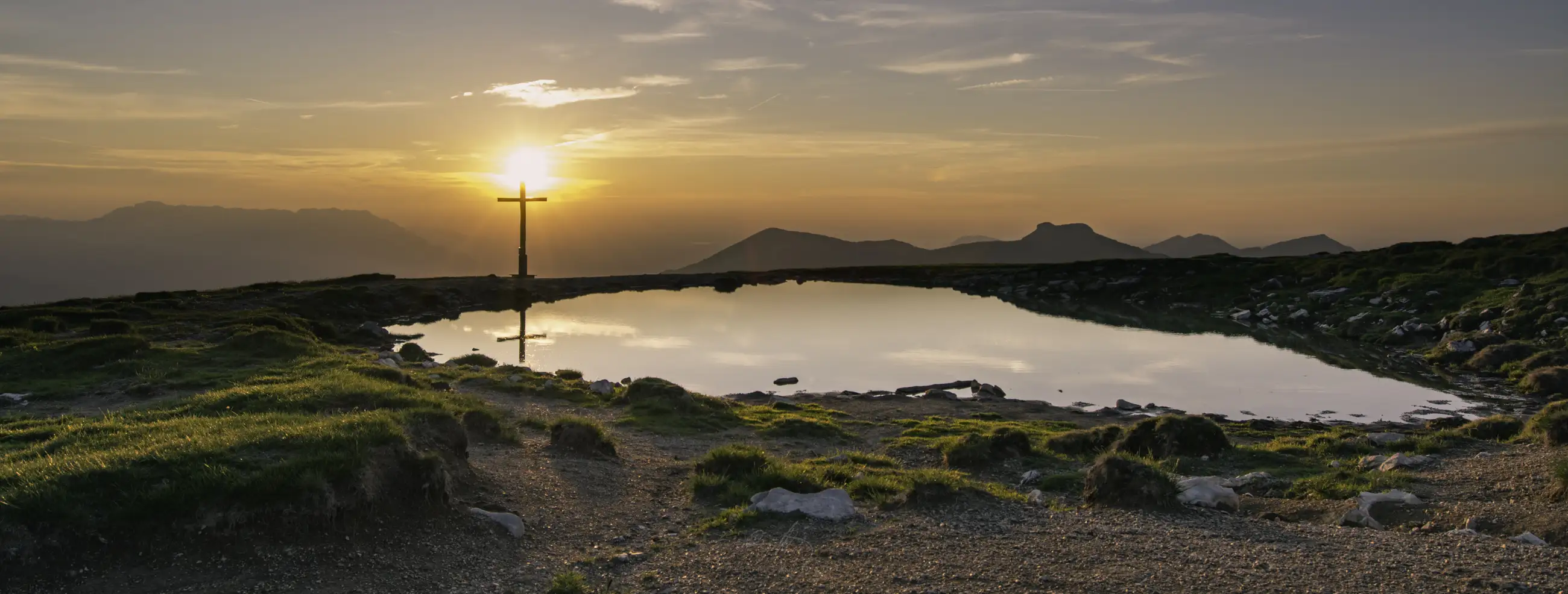 A tranquil mountain pond reflects a colorful sunset sky and a lone cross standing on the rocky shore, with distant mountains visible in the background.