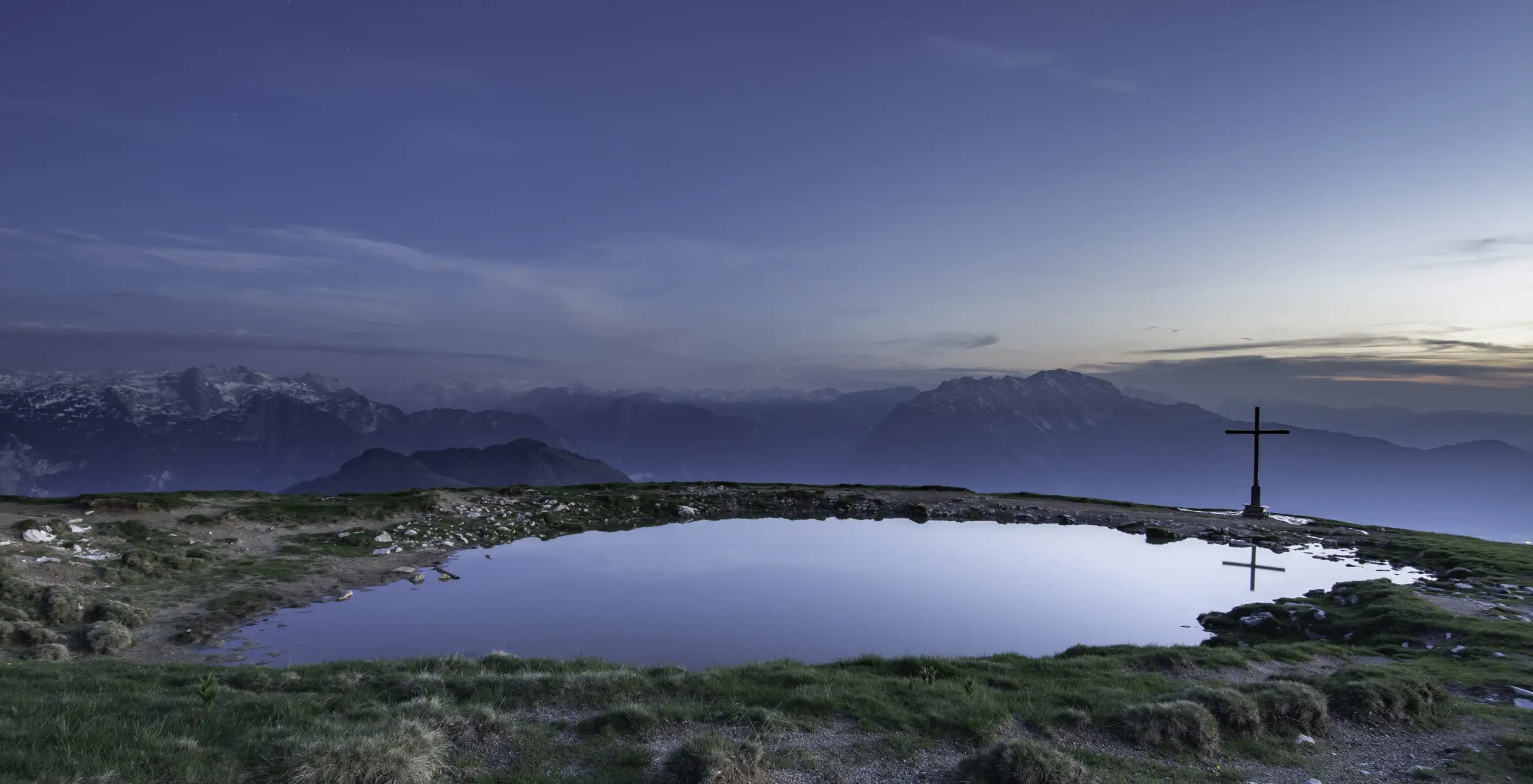 A grassy mountain plateau with a pond, scattered rocks, and a large cross on the right. Snow-capped mountains and a hazy sky appear in the distance.