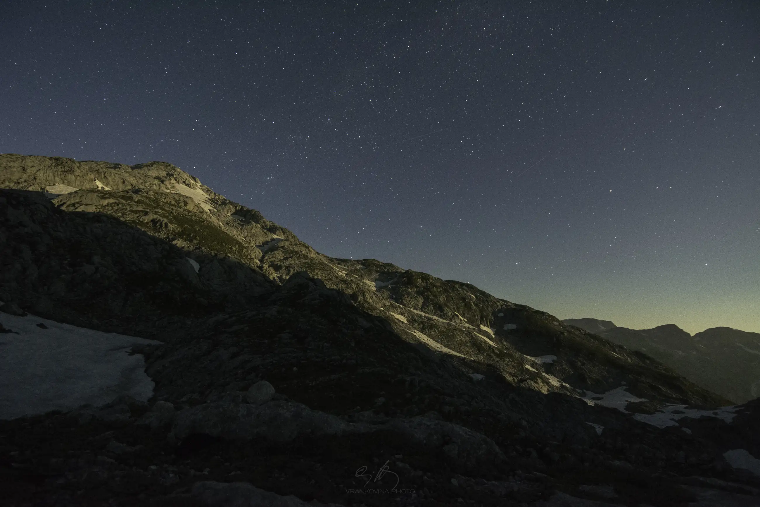 A rocky mountain slope is illuminated under a starry night sky, with patches of snow visible on the ground and distant mountains fading into the horizon.