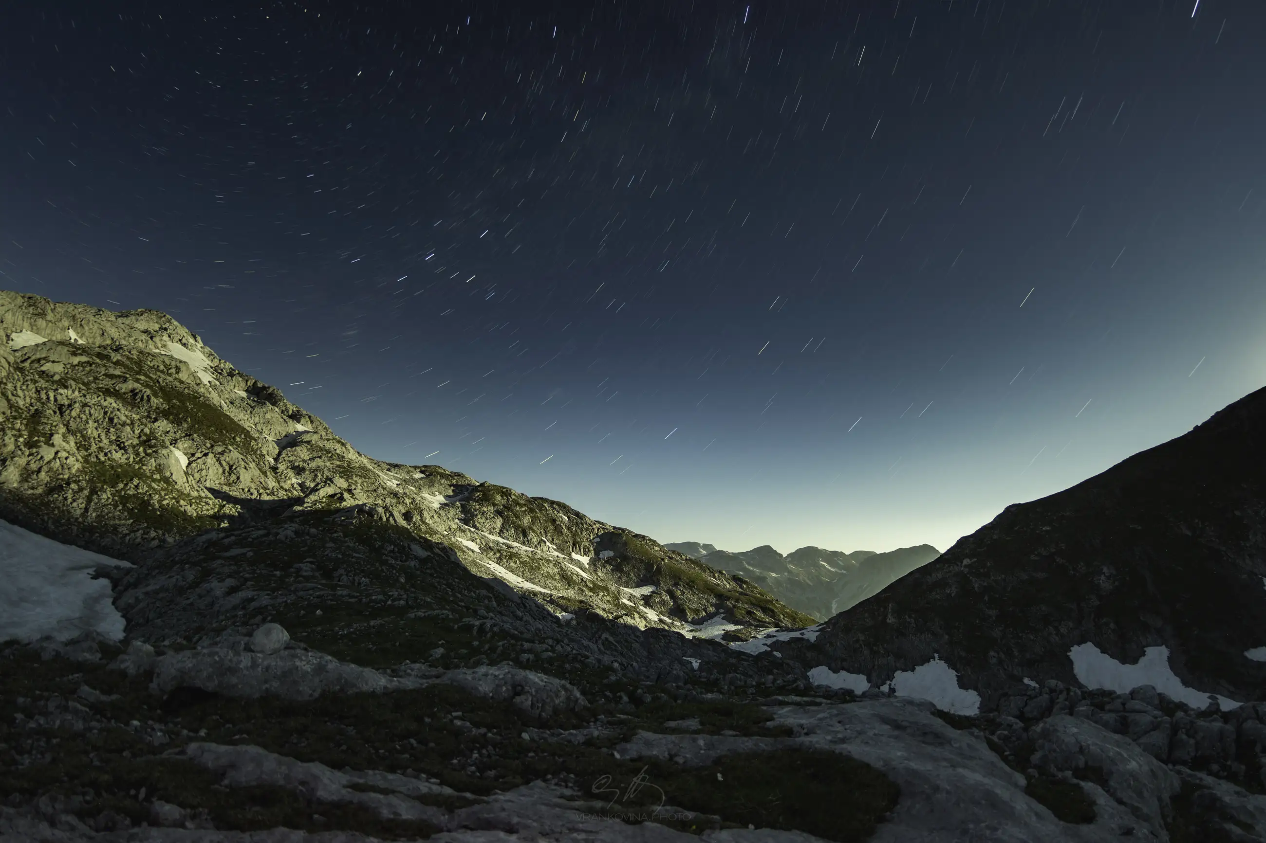 Rocky mountain landscape at night under a starry sky, with star trails visible. The slopes are partially covered in snow, and distant mountain peaks are seen under a clear, dark blue sky.