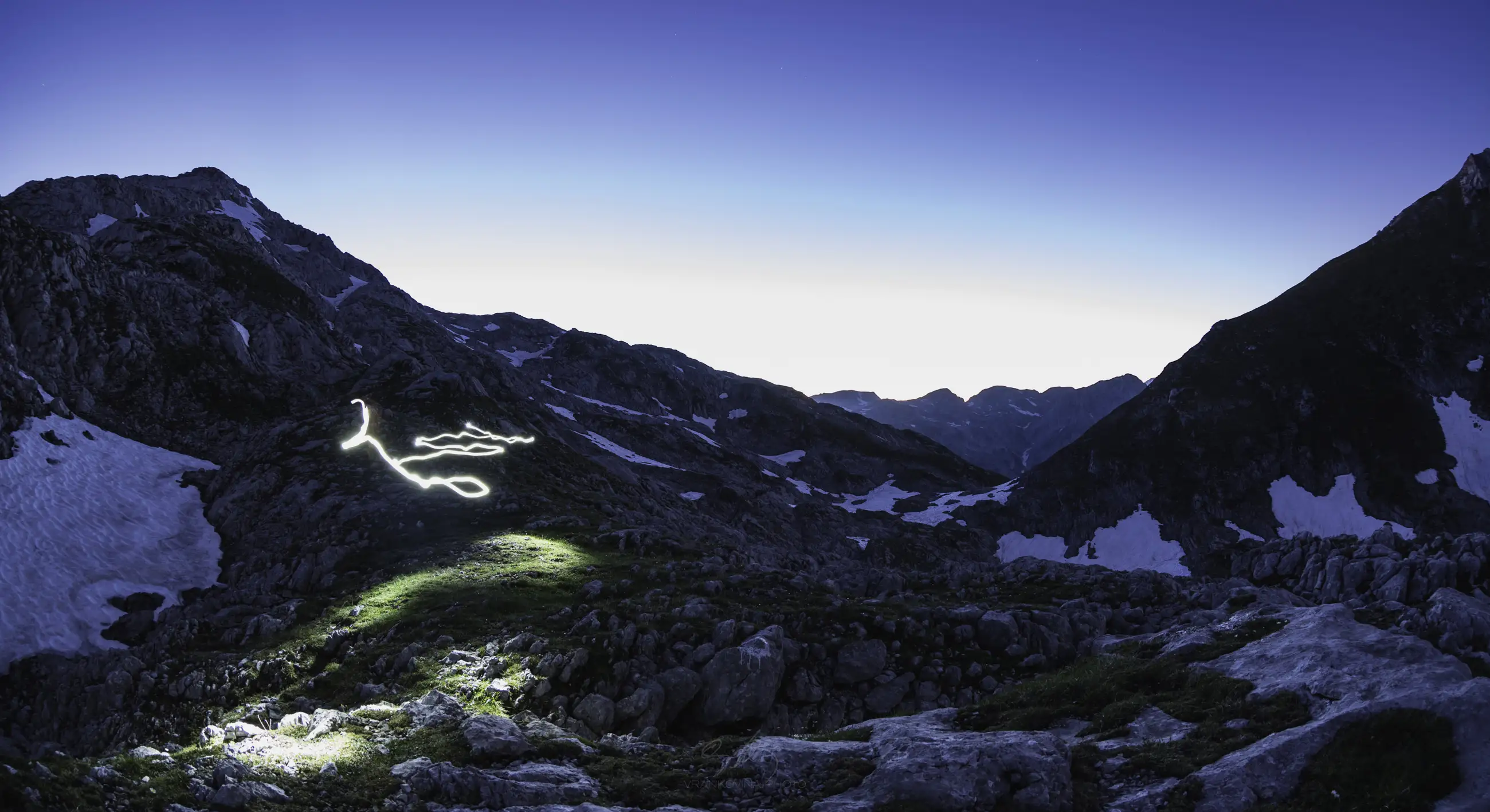 A mountain landscape at dusk with patches of snow. The sky is clear, and a glowing, swirling light trail appears on a grassy slope in the foreground, creating a surreal effect.