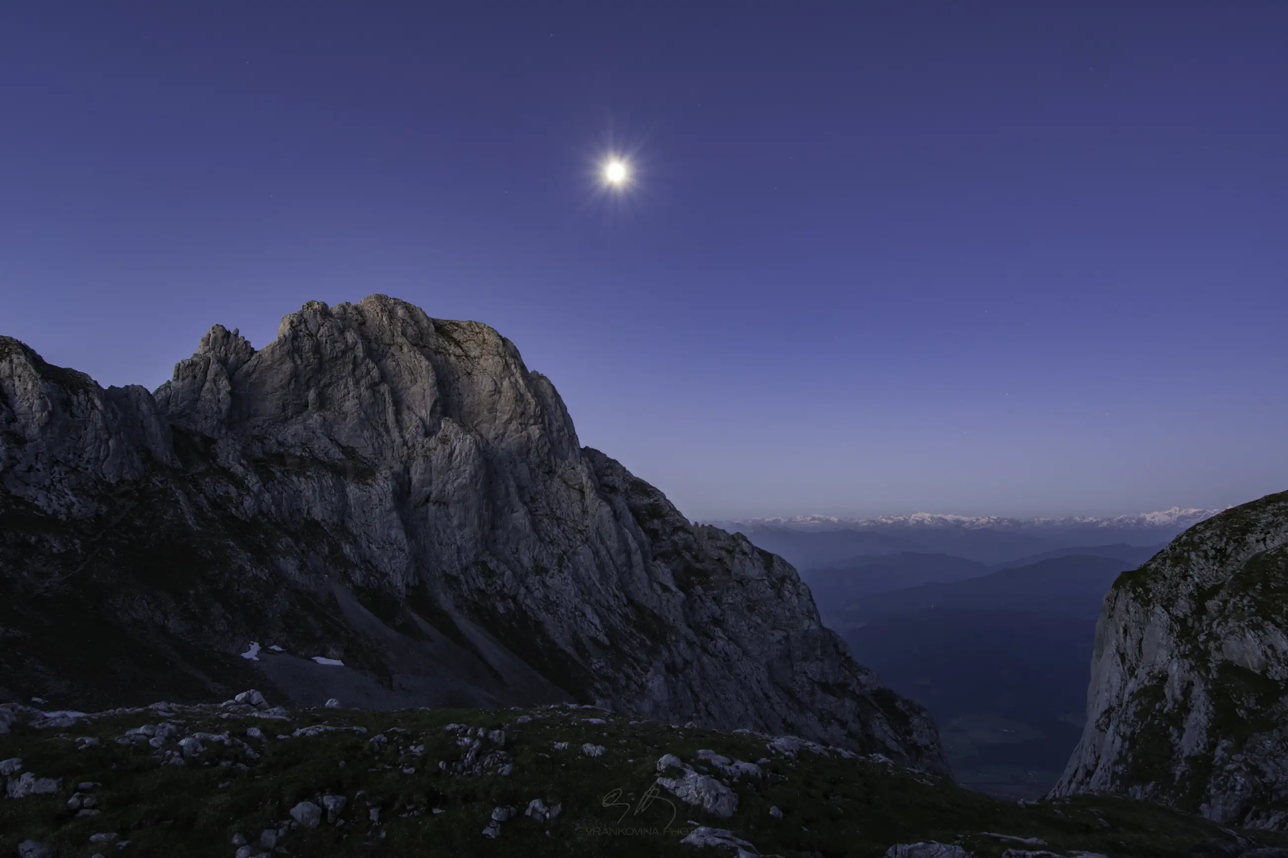 A rocky mountain peak under a clear twilight sky with the moon shining above; distant snow-capped mountains are visible on the horizon.
