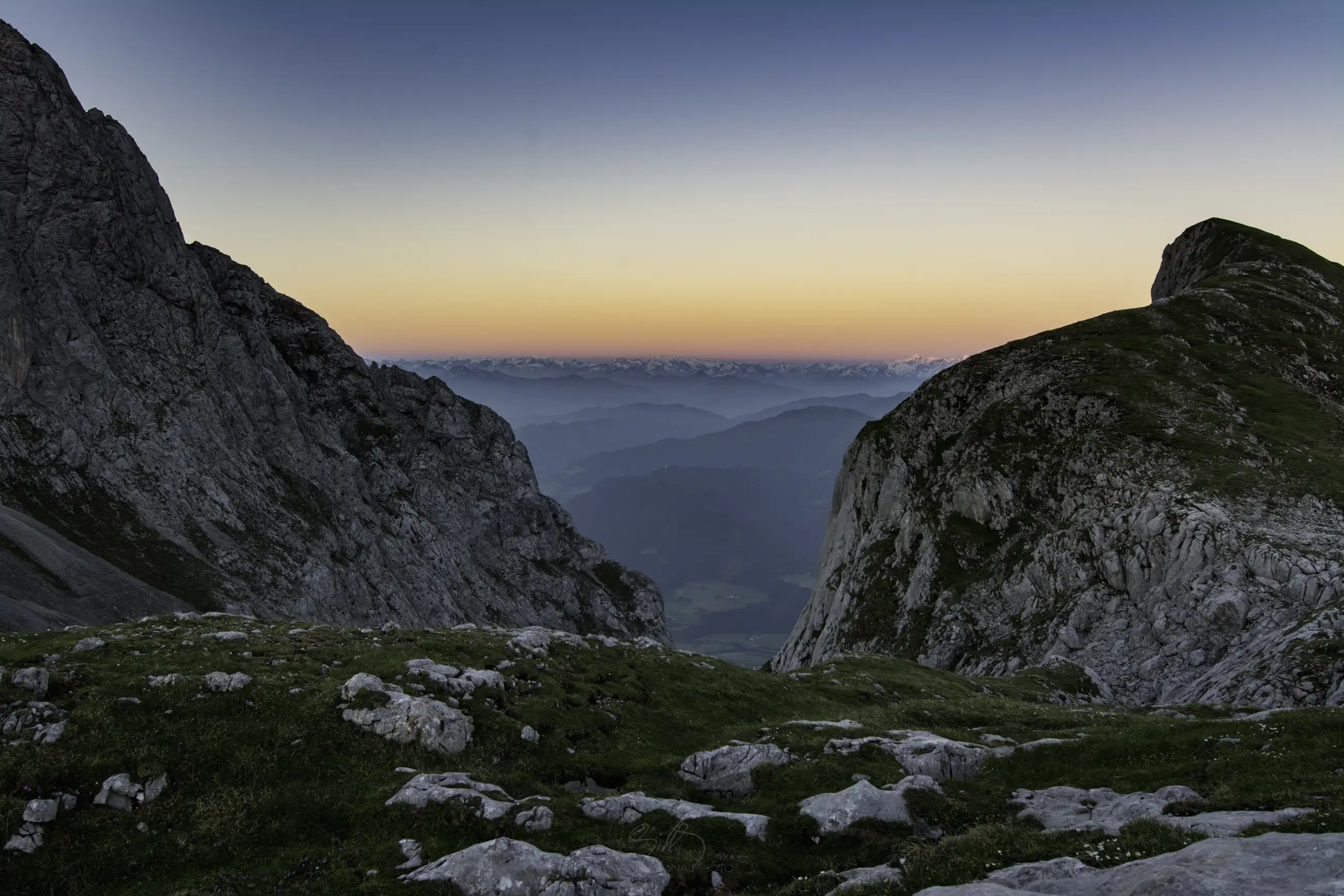 A mountain pass with rocky cliffs on both sides and grassy terrain in the foreground, overlooking distant mountains under a colorful sunrise or sunset sky.