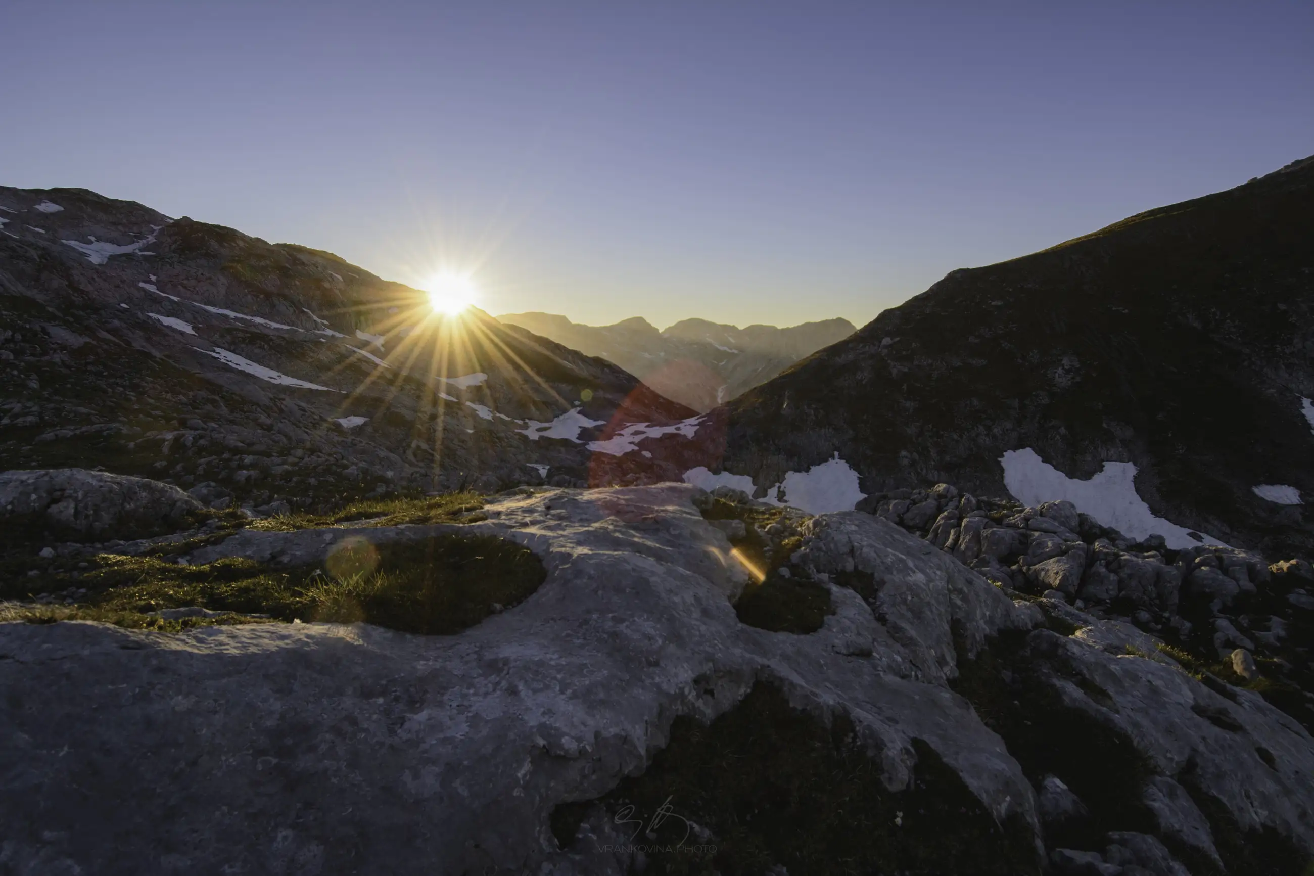 The sun sets behind distant mountains, casting a warm glow over rocky terrain with patches of snow and sparse grass under a clear blue sky.
