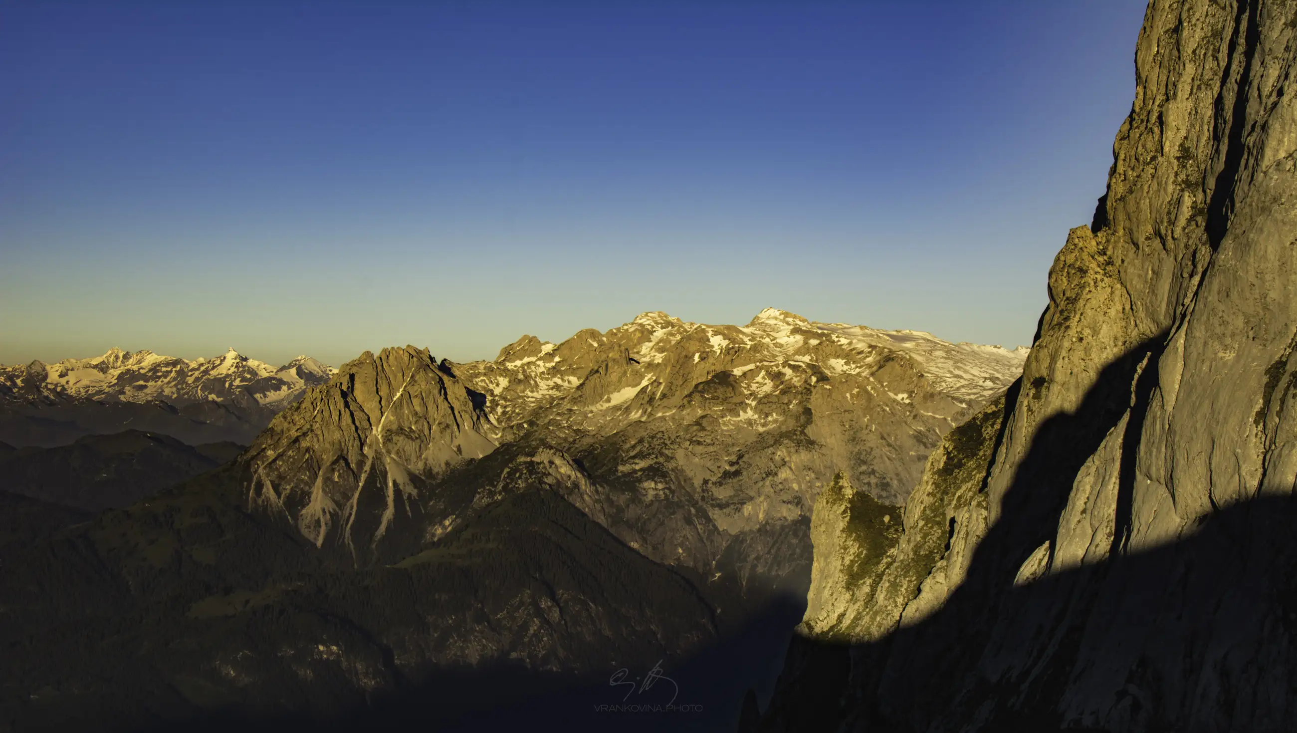 Mountain range with rugged peaks and patches of snow, bathed in warm sunlight under a clear blue sky; deep shadows fall on the rocky slopes in the foreground.