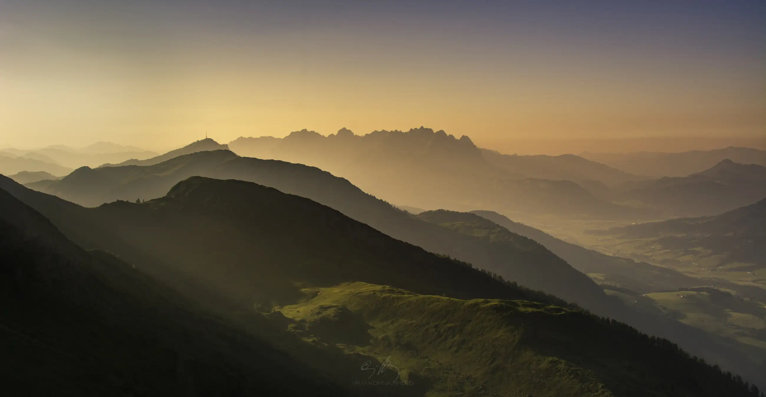Mountain ridges bathed in golden sunlight, with layers of mist creating a dreamy atmosphere; soft green slopes in the foreground and distant peaks fading into a pastel orange and blue sky at sunrise or sunset.