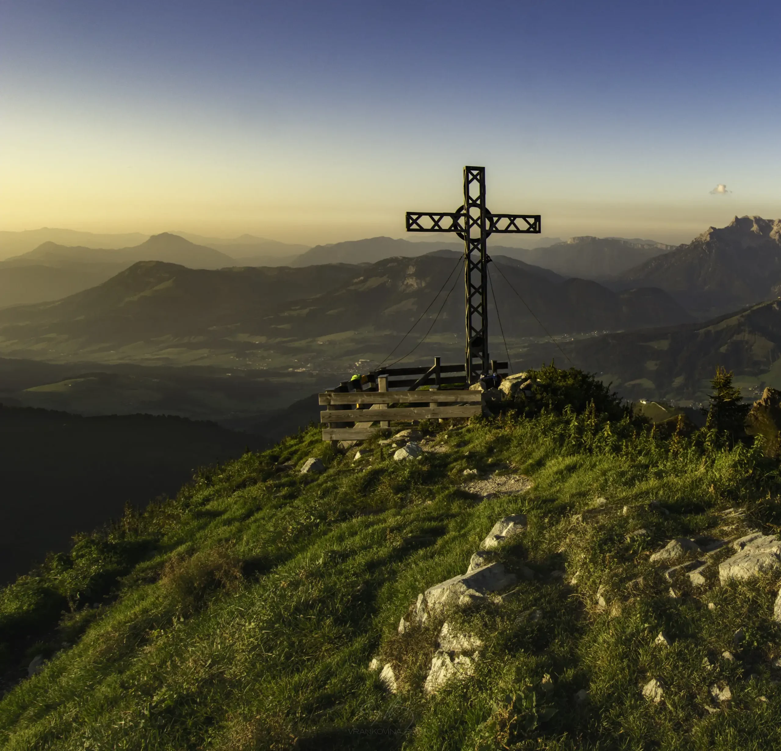 A large metal cross stands on a grassy mountaintop at sunset, overlooking a vast valley and distant mountains under a clear sky.