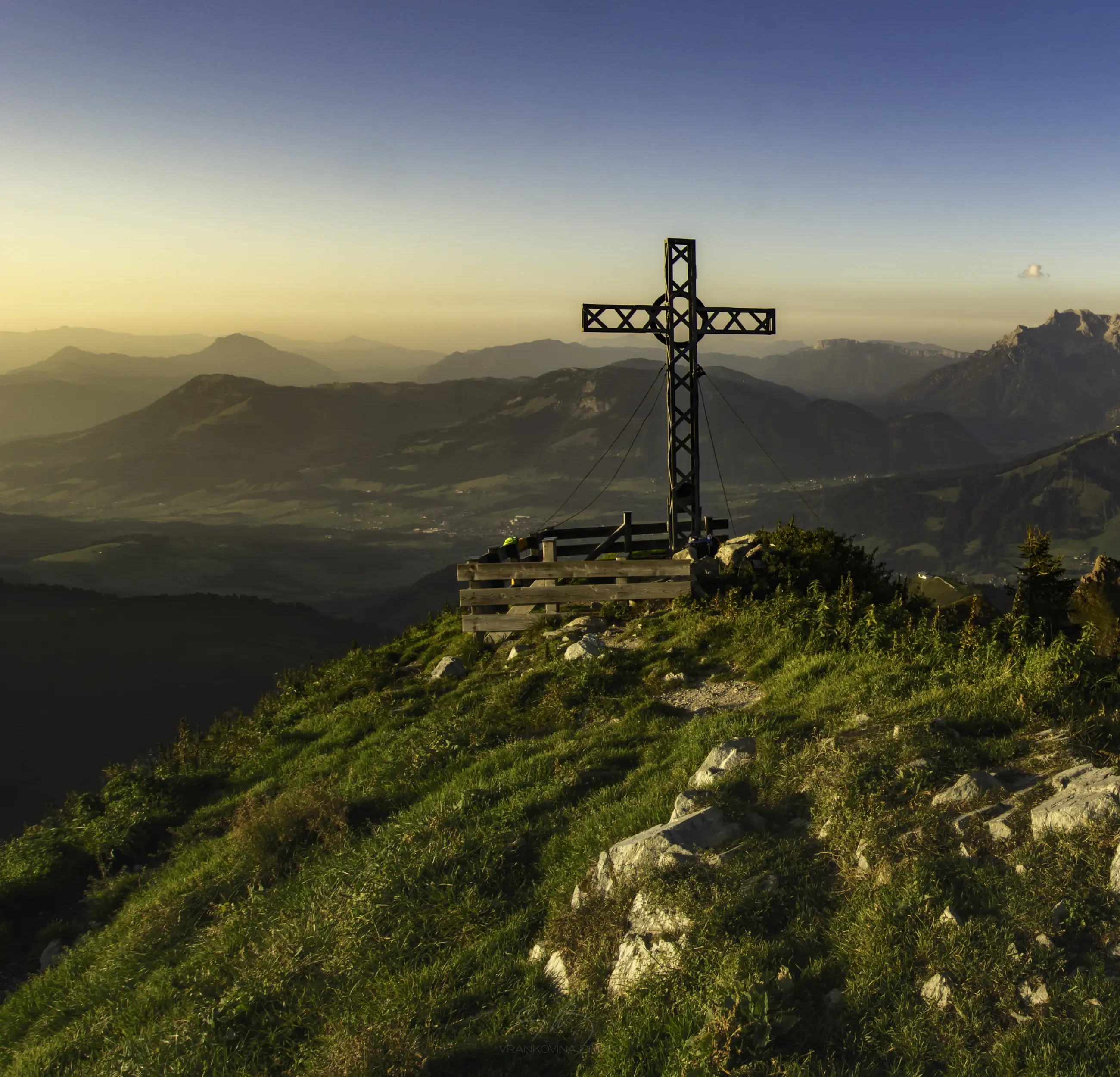 A large metal cross stands on a grassy mountaintop at sunset, overlooking a vast valley and distant mountains under a clear sky.