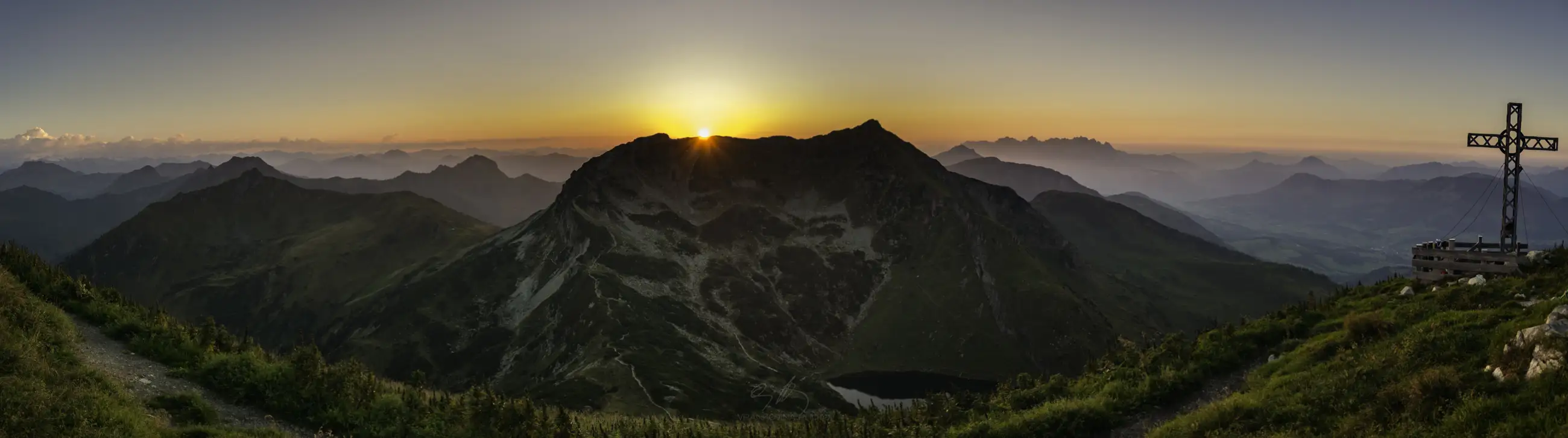 Panoramic view of a mountain range at sunrise, with the sun peeking over a central peak, grassy slopes in the foreground, and a large cross on the right side near a small building.