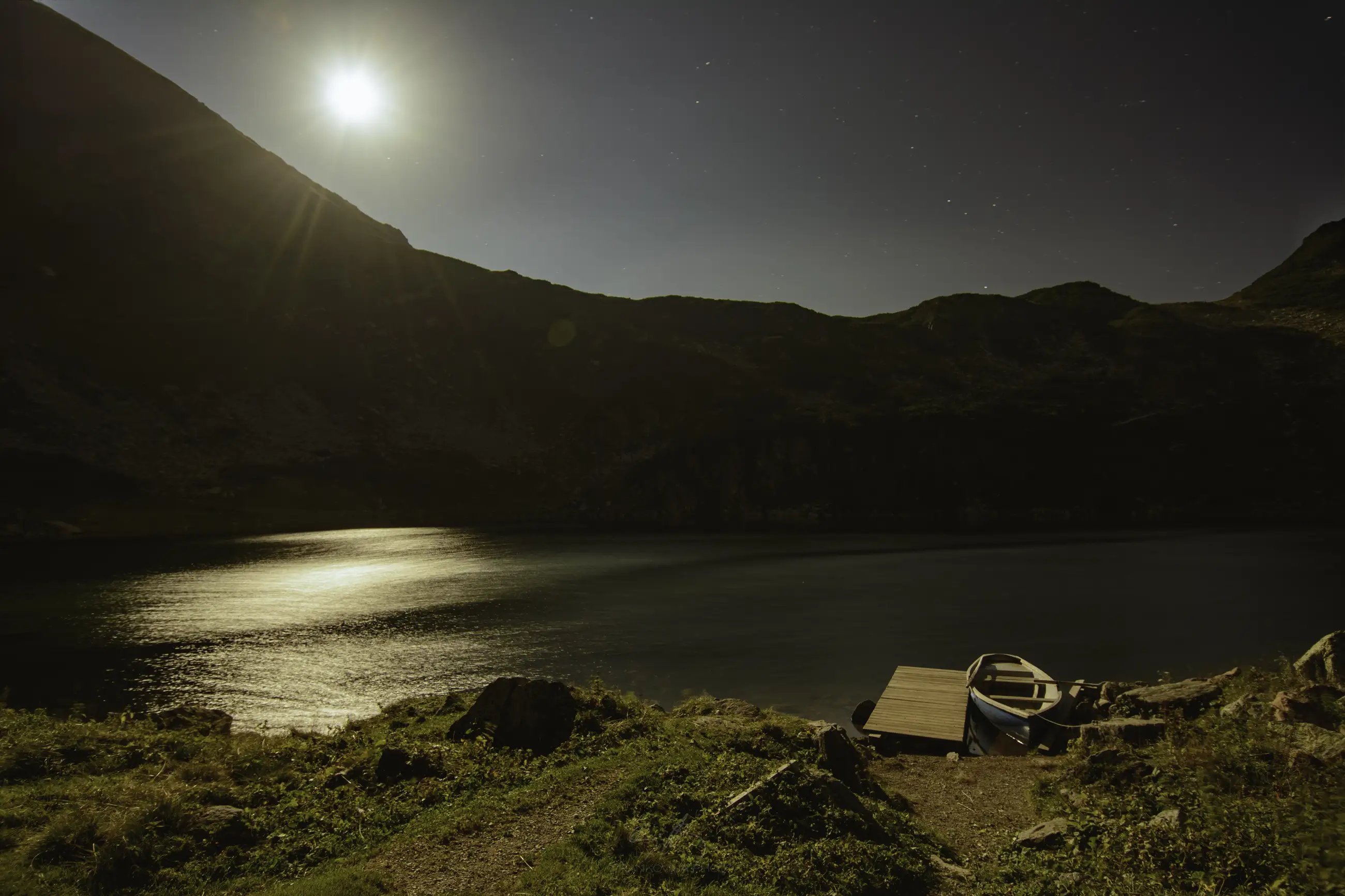 A small boat is tied to a wooden dock beside a calm lake at night, with moonlight shining over the water and mountains silhouetted in the background.
