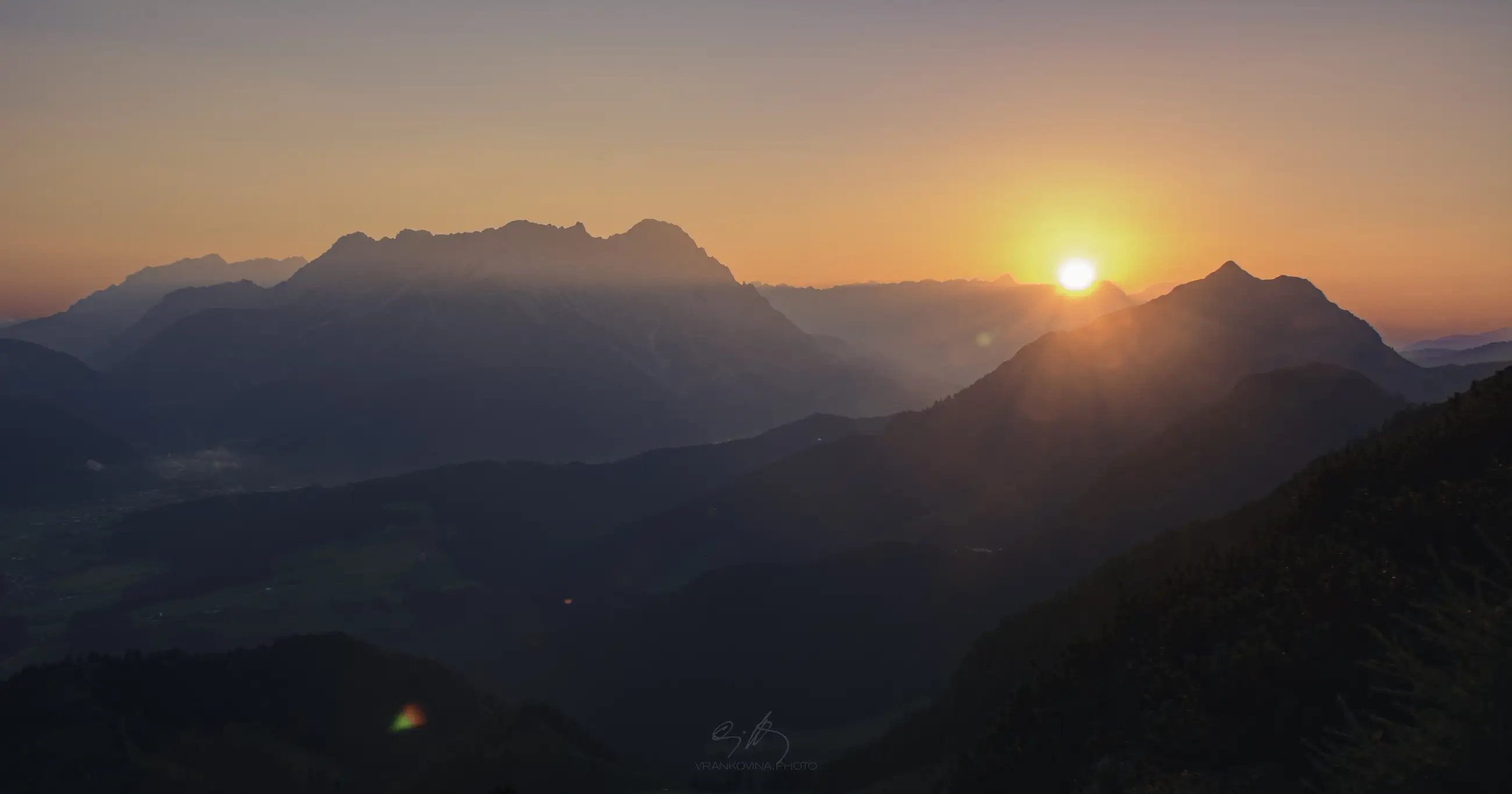 Sunrise over a mountain range, with the sun partially visible above the peaks, casting a warm orange and pink glow across the sky and misty blue valleys below.