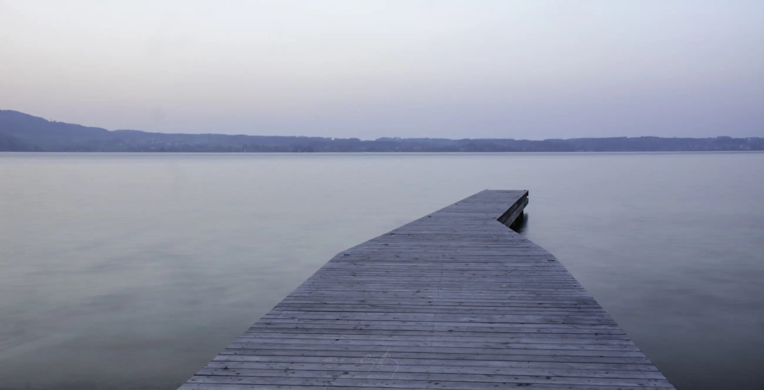 A wooden dock extends out over calm, still water under a soft, pale sky at dusk. Distant hills line the horizon, and the peaceful scene is devoid of people or boats.