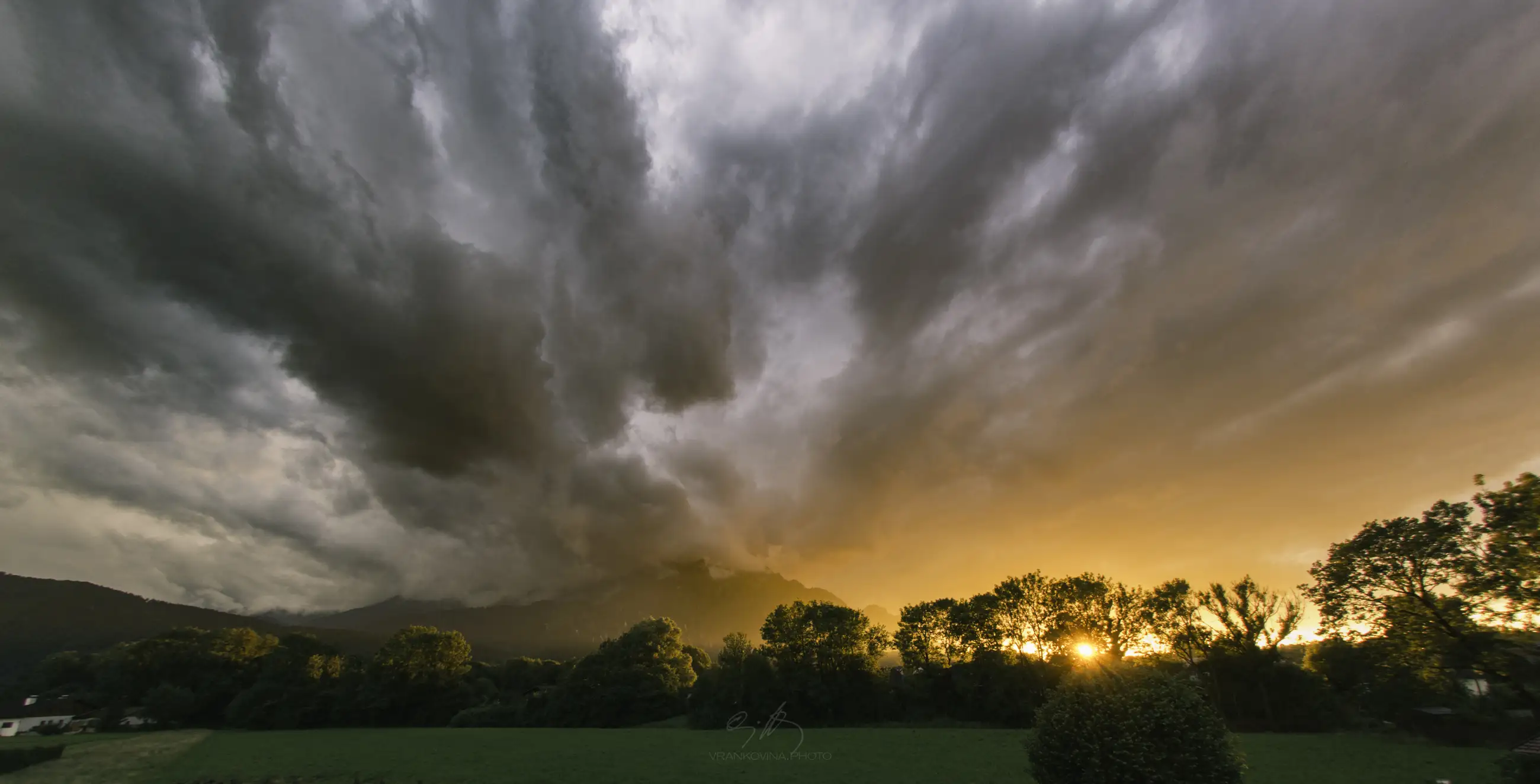 Fiery orange sunset illuminates clouds above a mountain partially obscured by smoke or mist, with dark trees and a green field in the foreground.