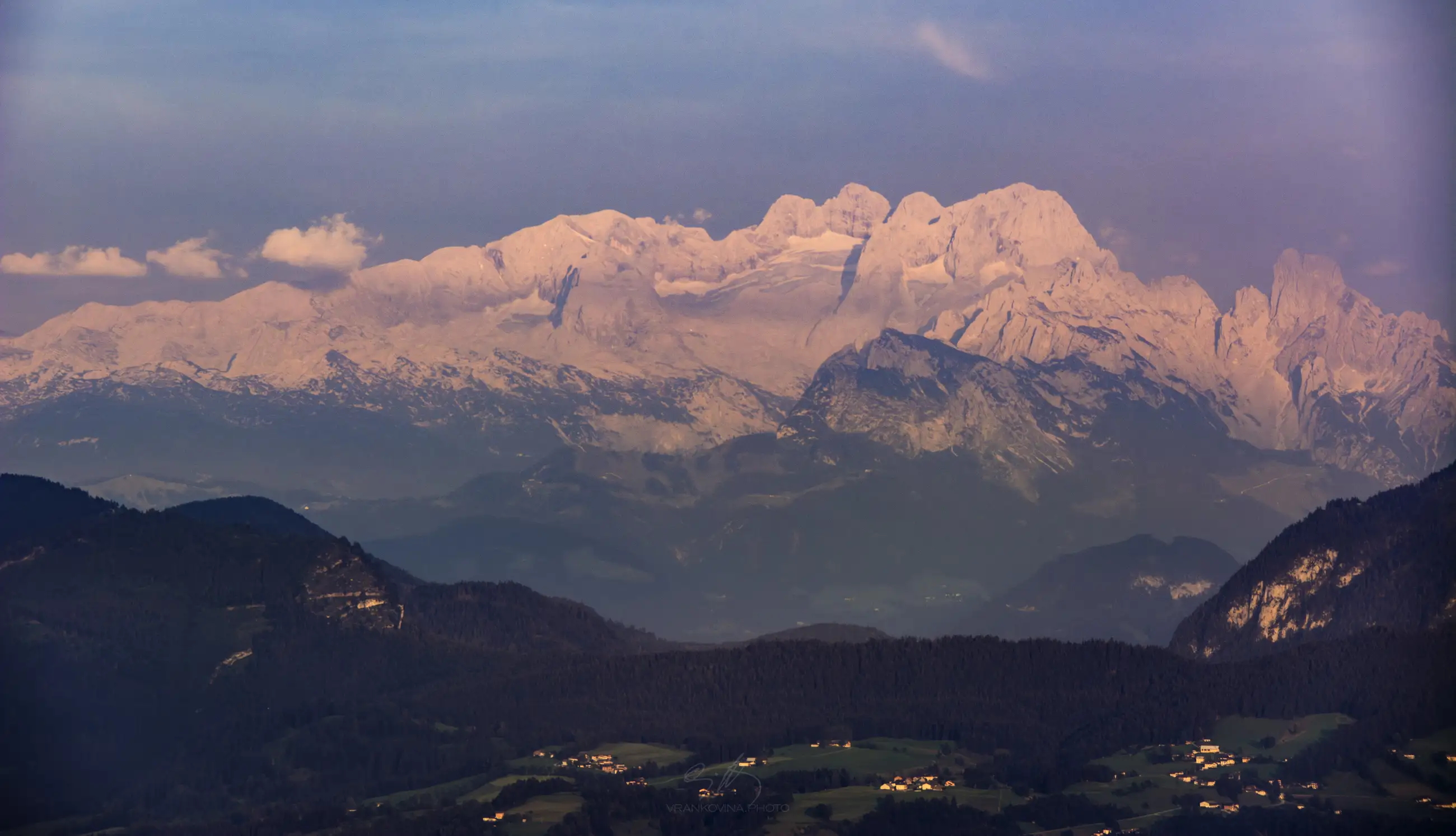 Snow-capped mountain peaks glow pink at sunset, with green hills, scattered houses, and forests below under a hazy sky.