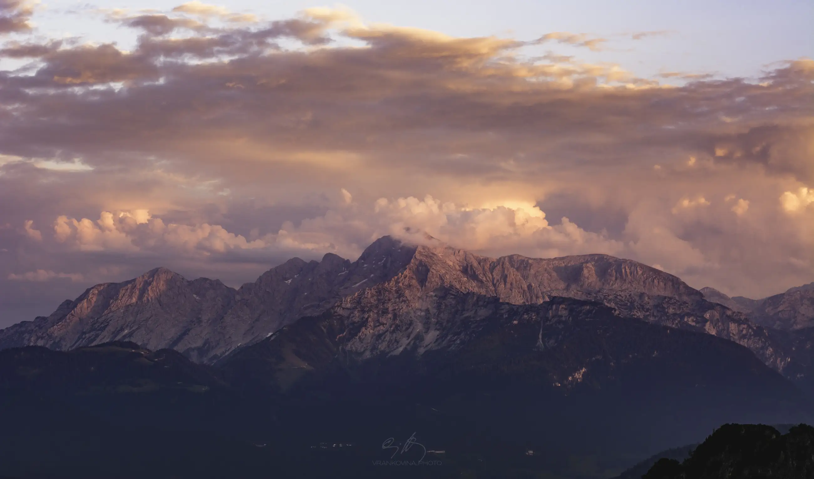 Mountain range under a colorful sky at sunset, with soft pink and orange clouds illuminating the rugged peaks. The mountains are partially covered in shadows, creating a dramatic and serene landscape.