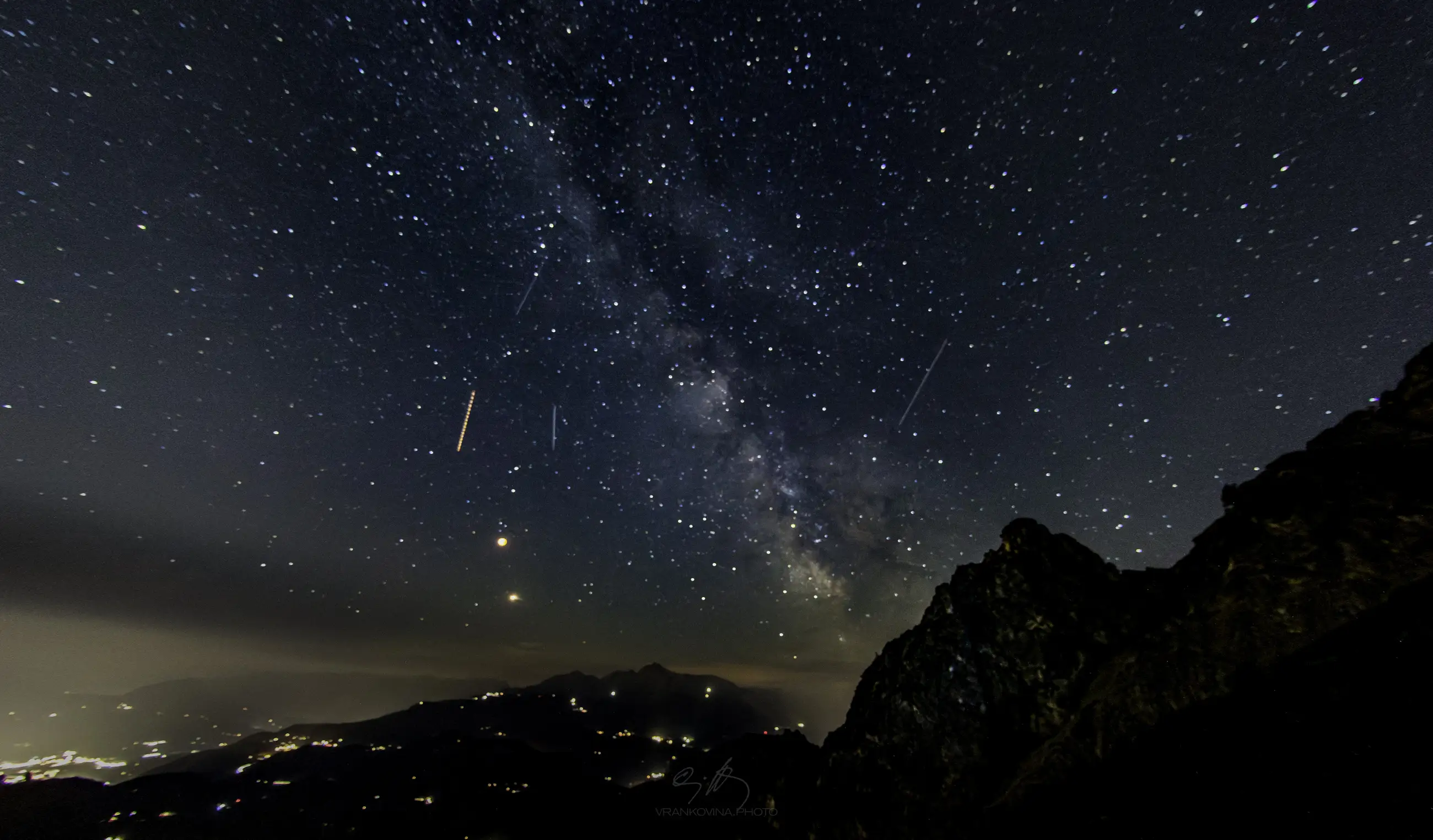 A clear night sky filled with stars and the Milky Way, plane tracks visible, above dark mountain silhouettes and distant city lights on the horizon.