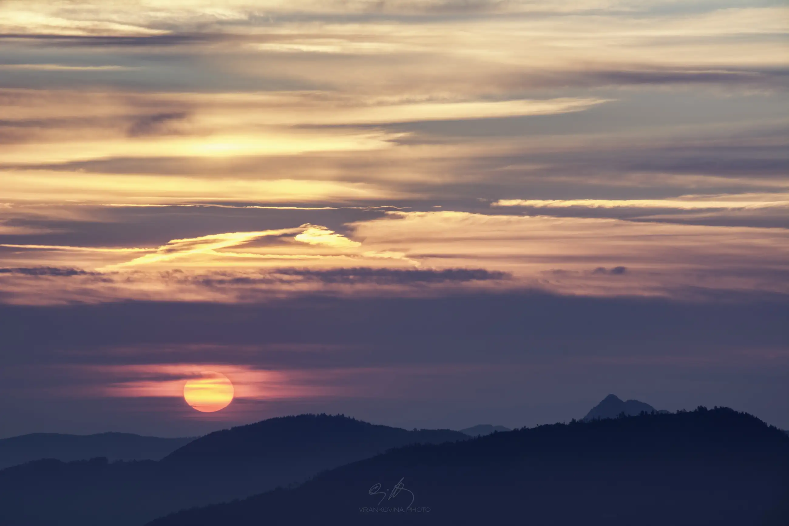 A colorful sunset over distant hills, with the sun low on the horizon and soft pink, orange, and purple clouds filling the sky above dark, silhouetted mountain ridges.