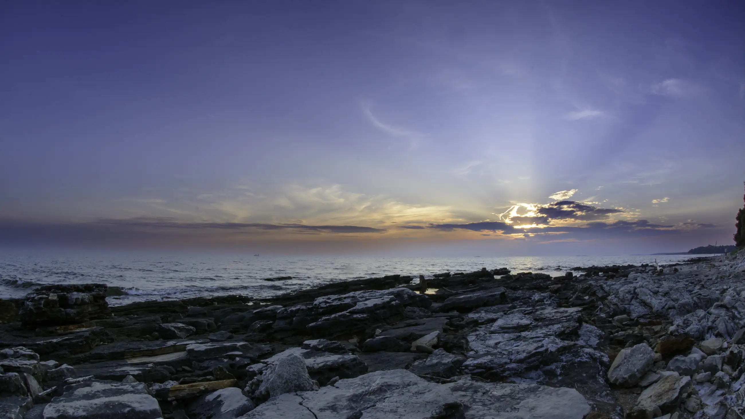 Rocky shoreline at sunset with the sun partially hidden behind clouds, casting a soft glow over the ocean. The sky transitions from blue to orange and purple, creating a serene and tranquil atmosphere.