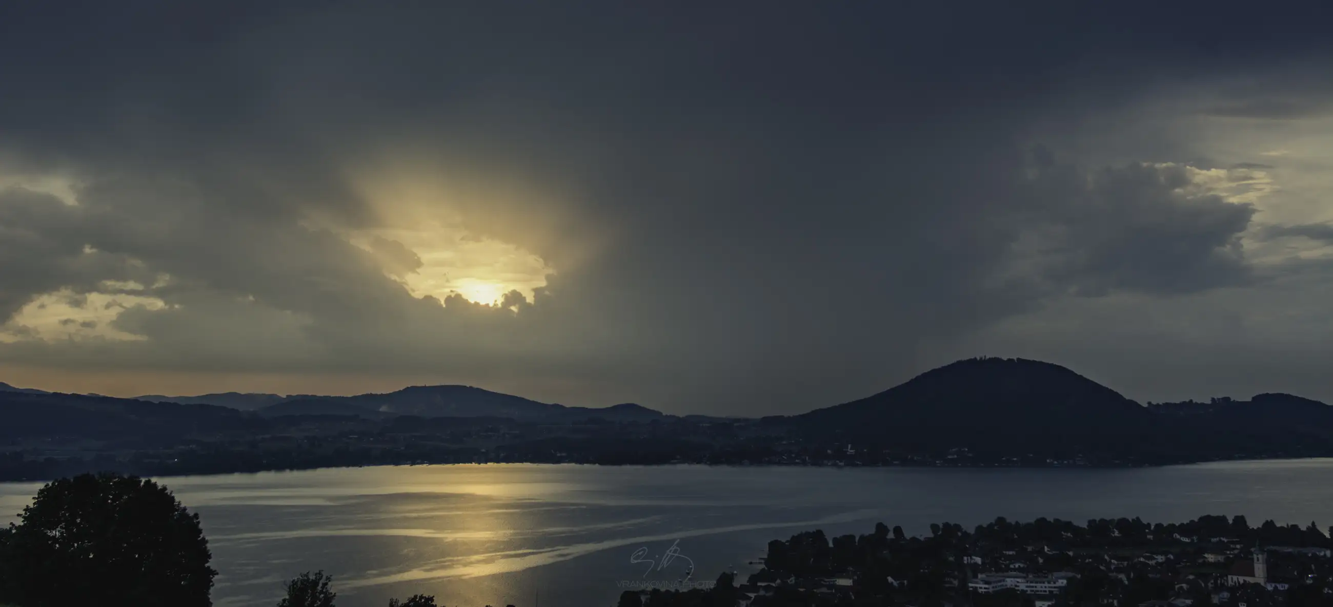 A dramatic sky with dark clouds and sunlight breaks over a calm lake, surrounded by hills and a small town along the shoreline. Trees and grass frame the foreground.