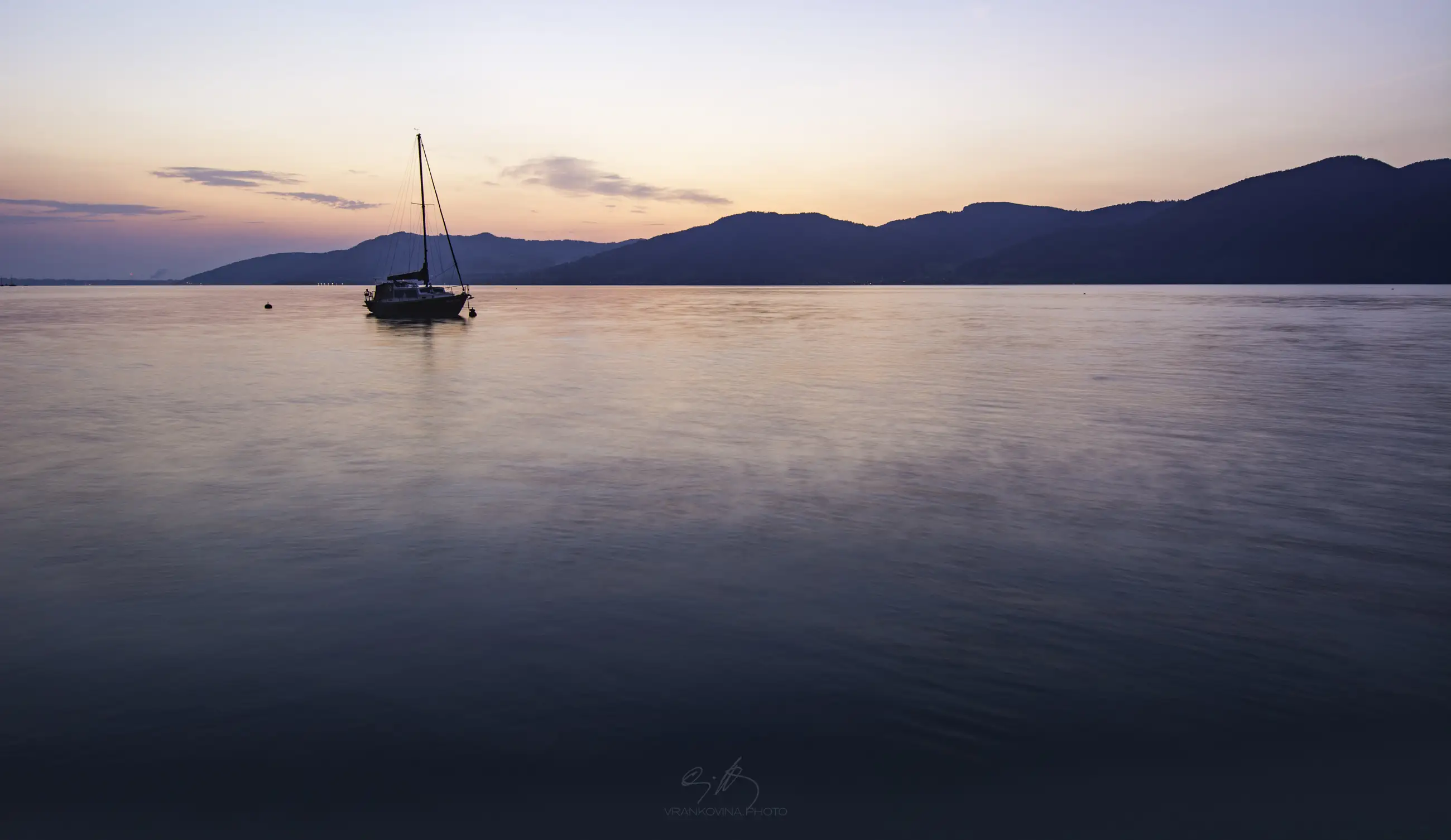 A sailboat floats on calm water at sunrise, with gentle ripples, silhouetted mountains, and a colorful sky with soft clouds in the background.