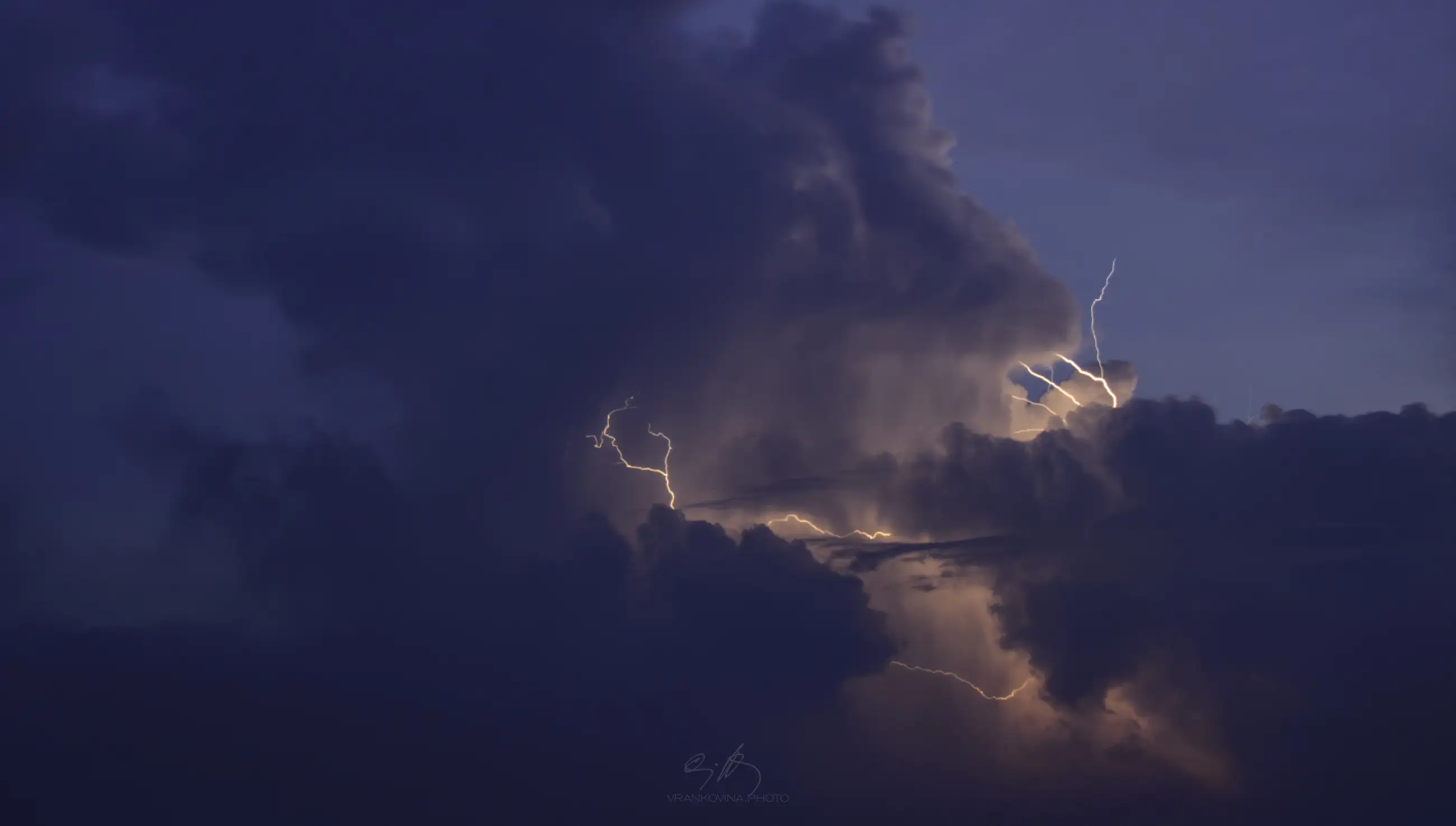 A dramatic sky filled with dark storm clouds, illuminated by flashes of lightning branching through the clouds during dusk or evening. The scene appears powerful and intense, with hints of orange and blue tones.