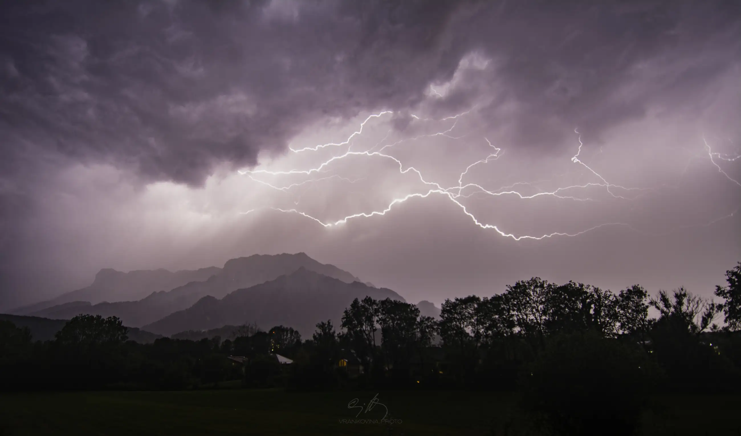 Bright lightning forks across a dark, cloudy night sky above silhouetted mountains and trees, illuminating the dramatic landscape below.
