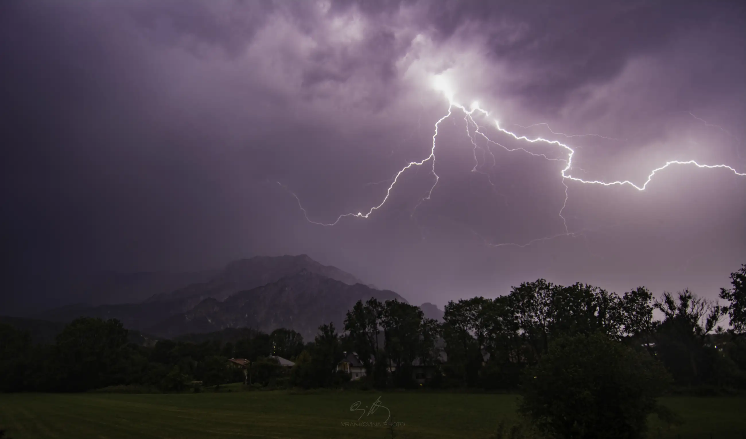 Bright lightning forks across a dark, cloudy night sky above silhouetted mountains and trees, illuminating the dramatic landscape below.