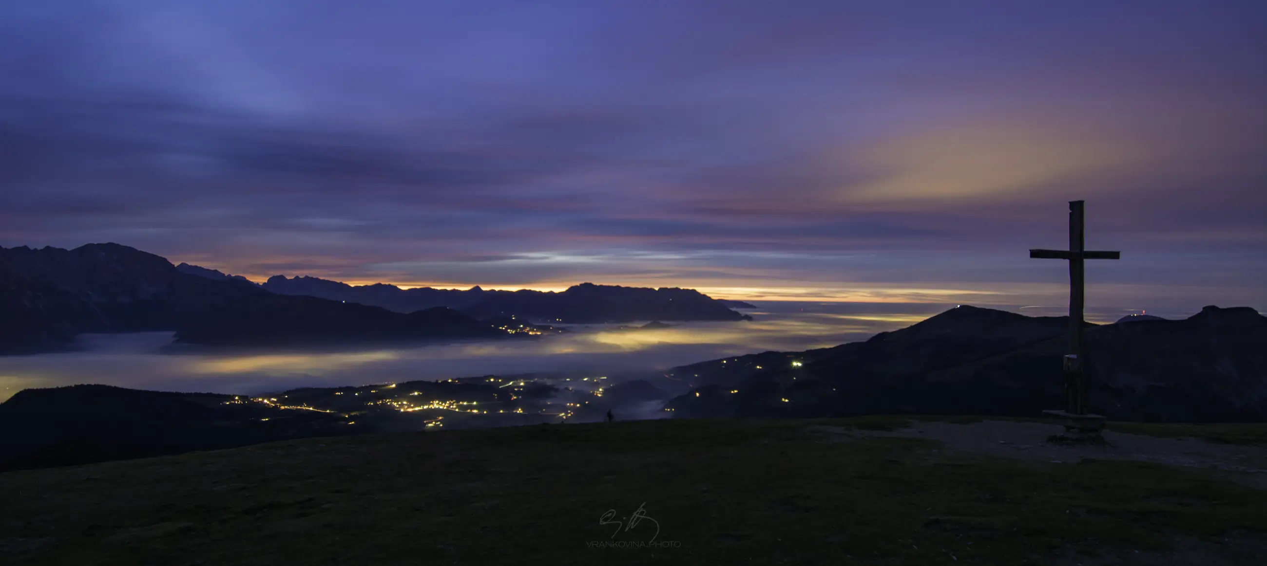 A twilight mountain landscape with glowing village lights below, mist covering the valleys, a cross on a hill in the foreground, and colorful clouds in a blue and pink sky over distant peaks.