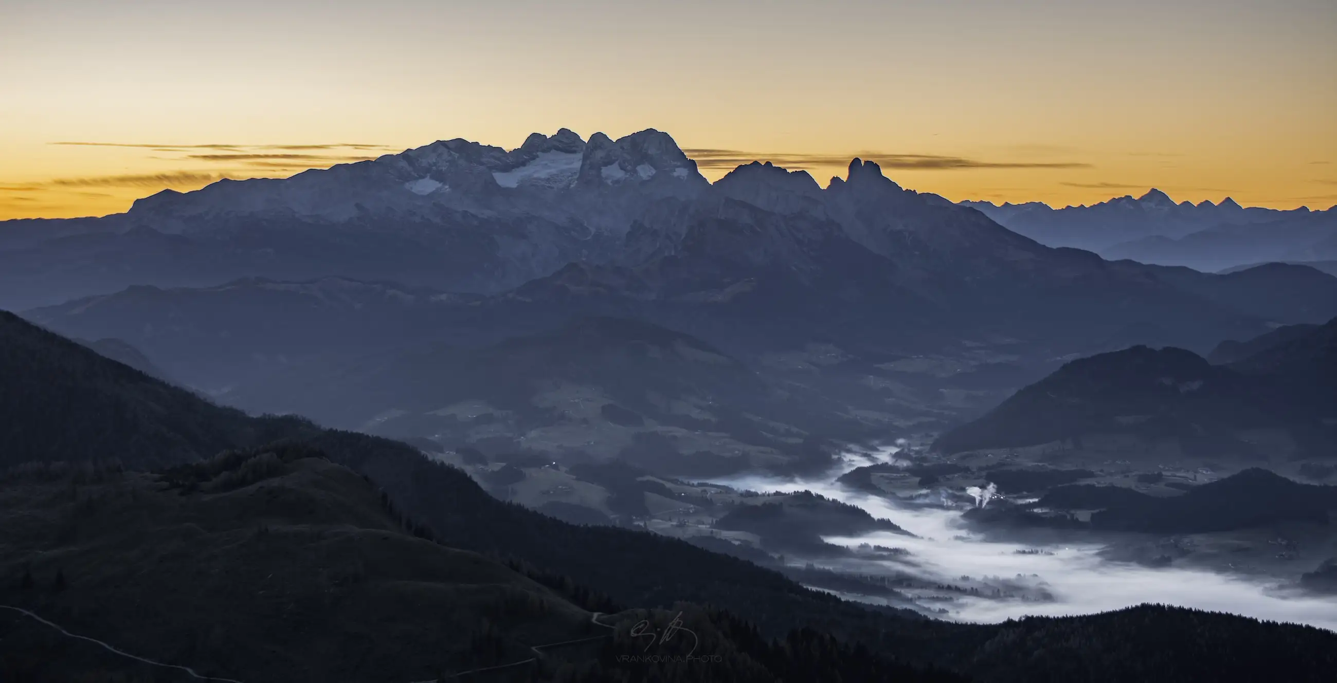 Sunrise over a mountain range with peaks silhouetted against an orange sky, rolling hills in the foreground, and valley clouds illuminated by early morning light.