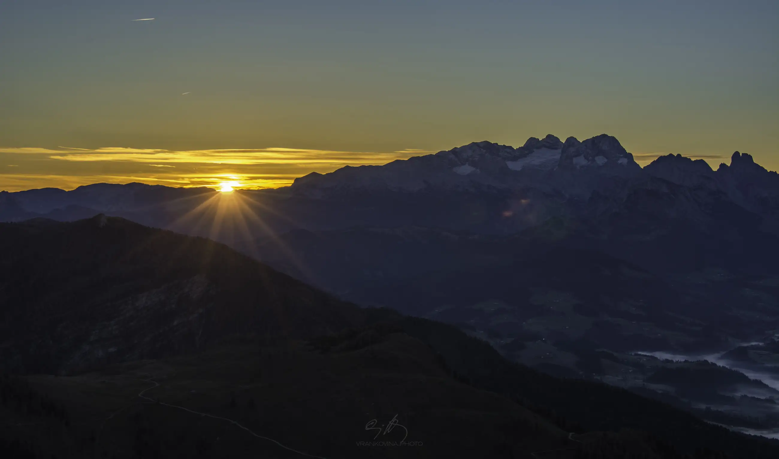Sunrise over mountain peaks, with the sun casting rays through a gap in the mountains, illuminating a valley below. The sky transitions from orange near the horizon to clear blue above.