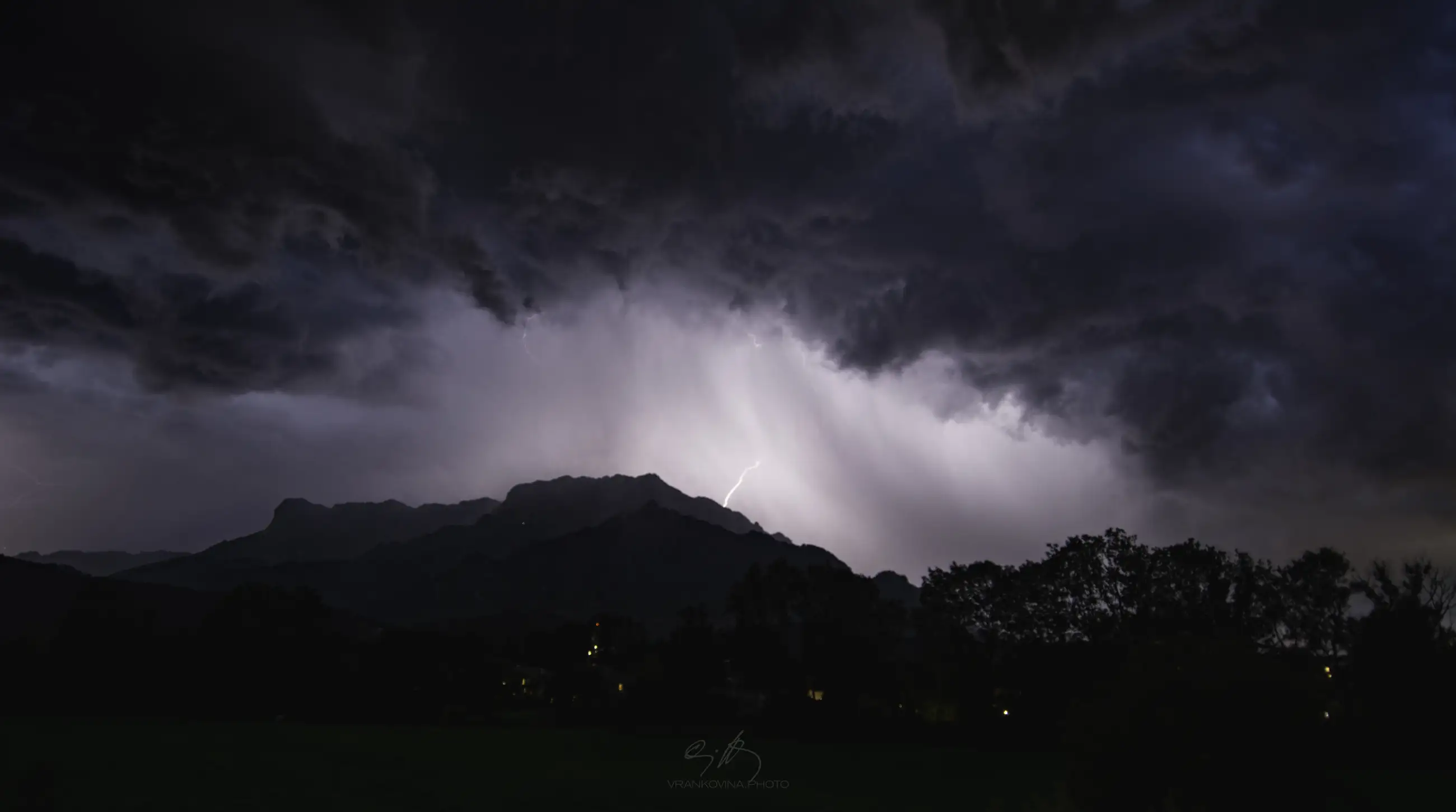 A dramatic night sky with dark clouds and a bright lightning bolt striking above silhouetted trees and distant mountains. The landscape is dimly lit under the stormy weather.