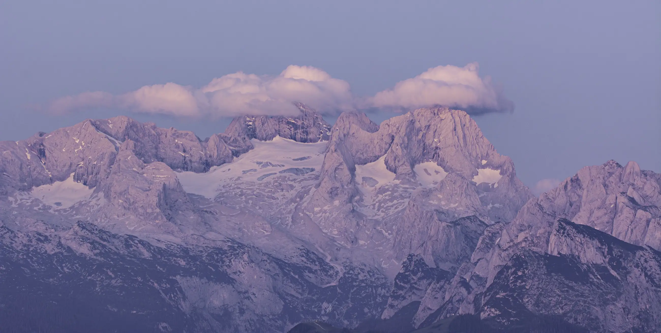 Snowy mountain peaks bathed in soft pink light at sunrise or sunset, with fluffy clouds floating above the summits against a pale blue sky.