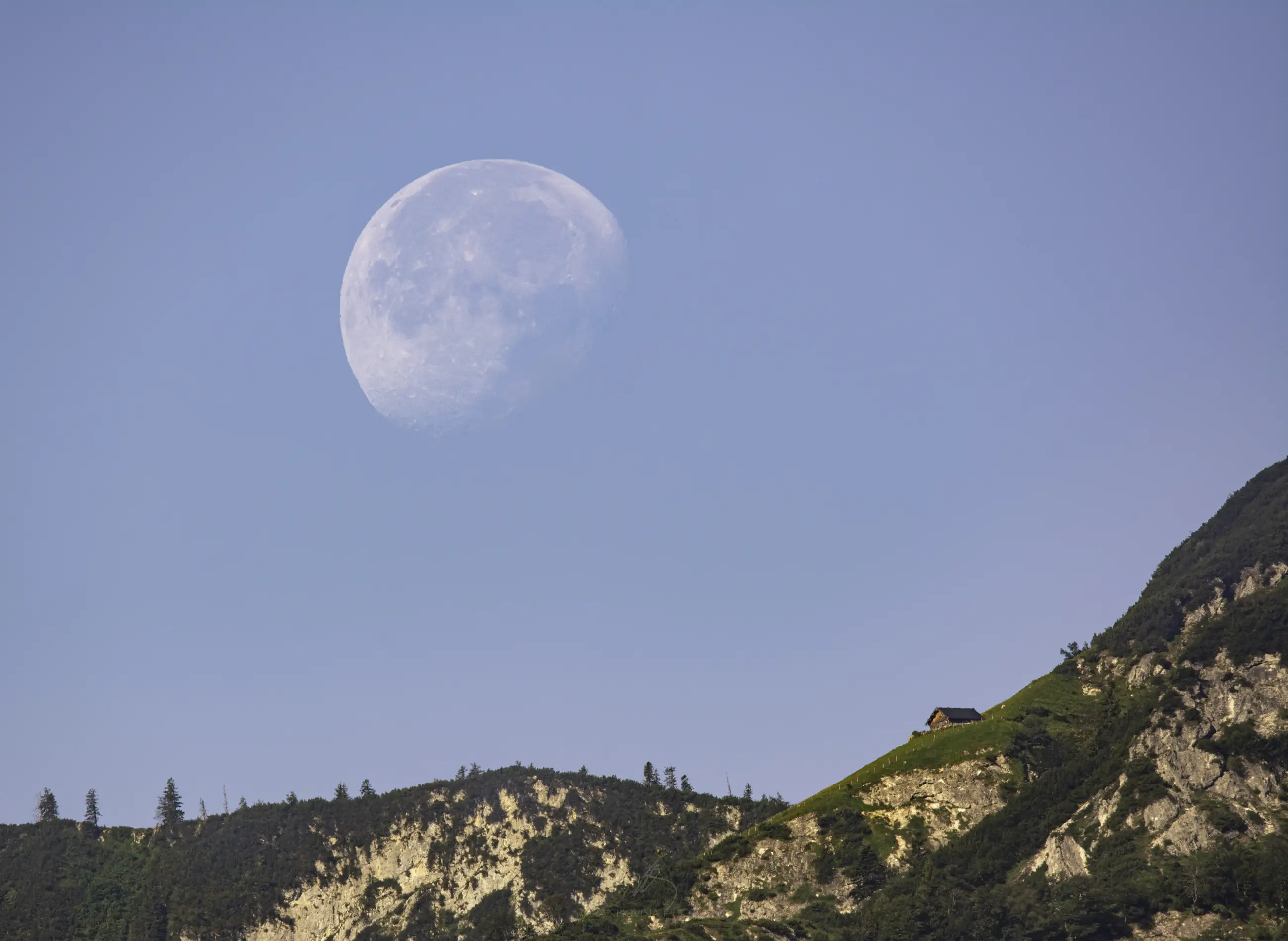 A bright, nearly full moon rises behind a steep, green mountain with rocky cliffs. A small house sits near the mountain&rsquo;s ridge under a clear blue sky.