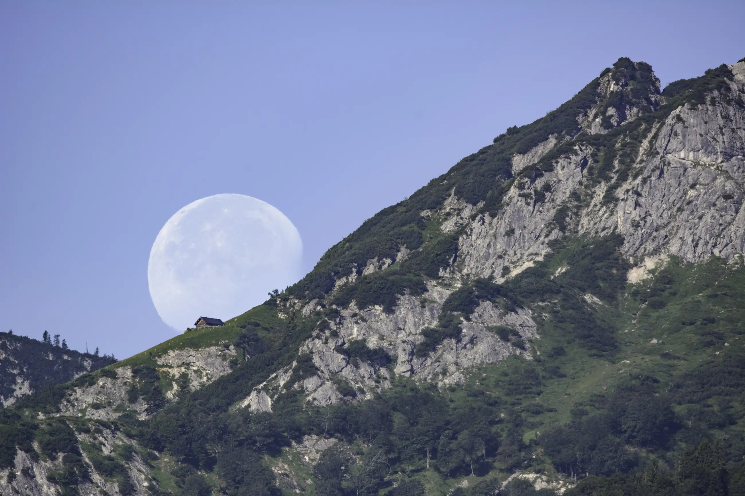 A bright, nearly full moon rises behind a steep, green mountain with rocky cliffs. A small house sits near the mountain&rsquo;s ridge under a clear blue sky.