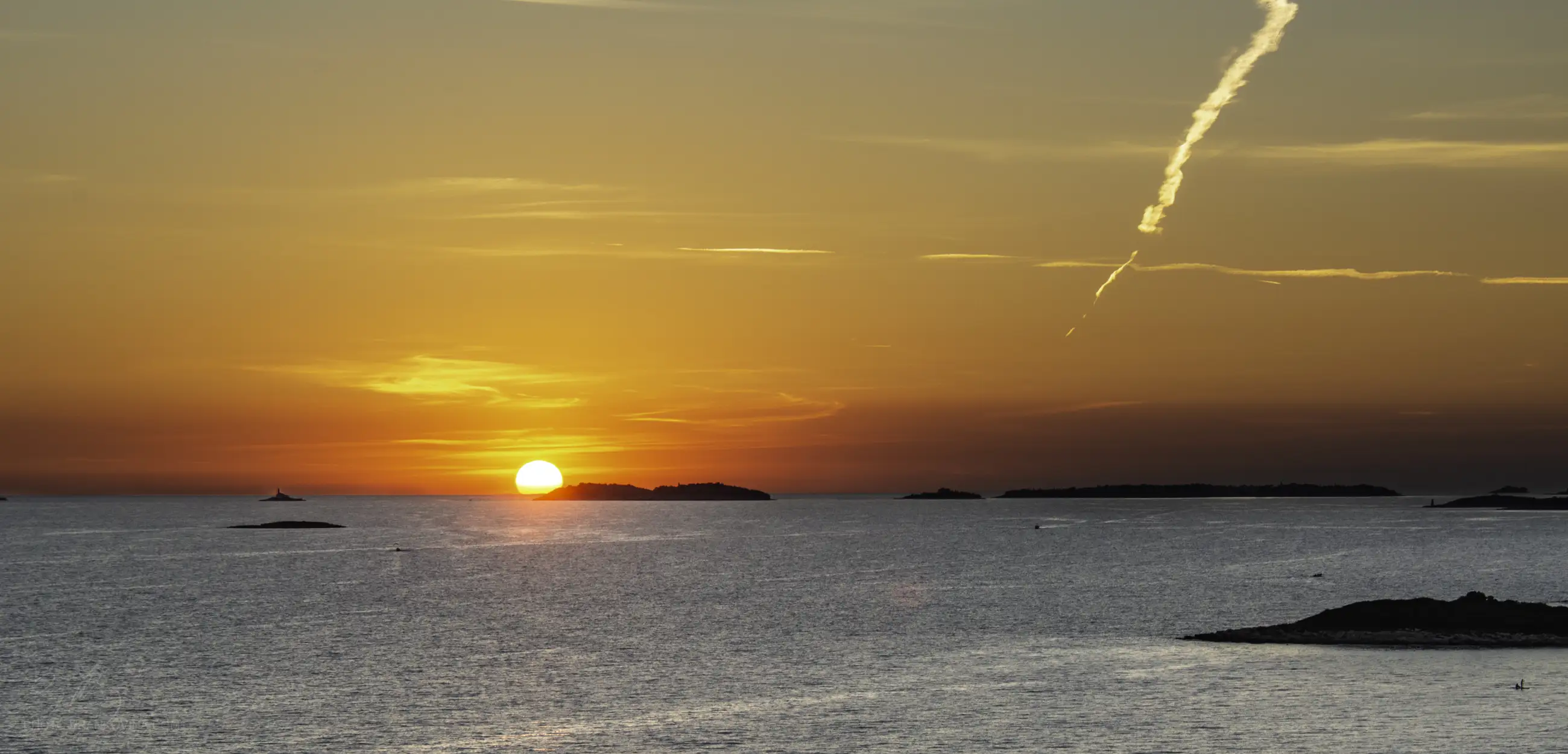 A vibrant sunset over a calm sea, with the sun partially visible near the horizon, small islands in the distance, and a streak of cloud or vapor trail diagonally across the orange and yellow sky.