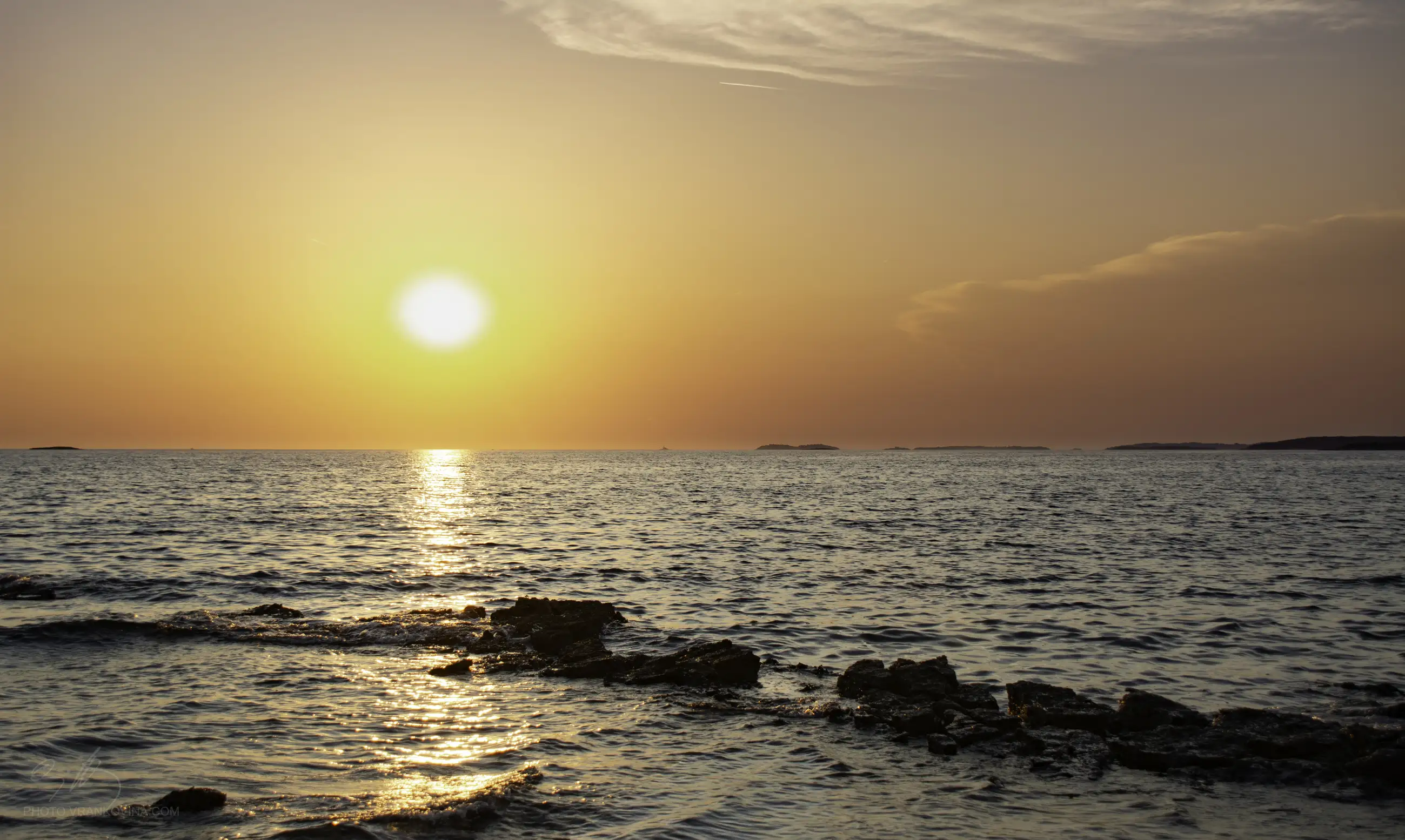 The sun sets over a calm ocean, casting an orange glow across the sky and water, with gentle waves and scattered rocks in the foreground.