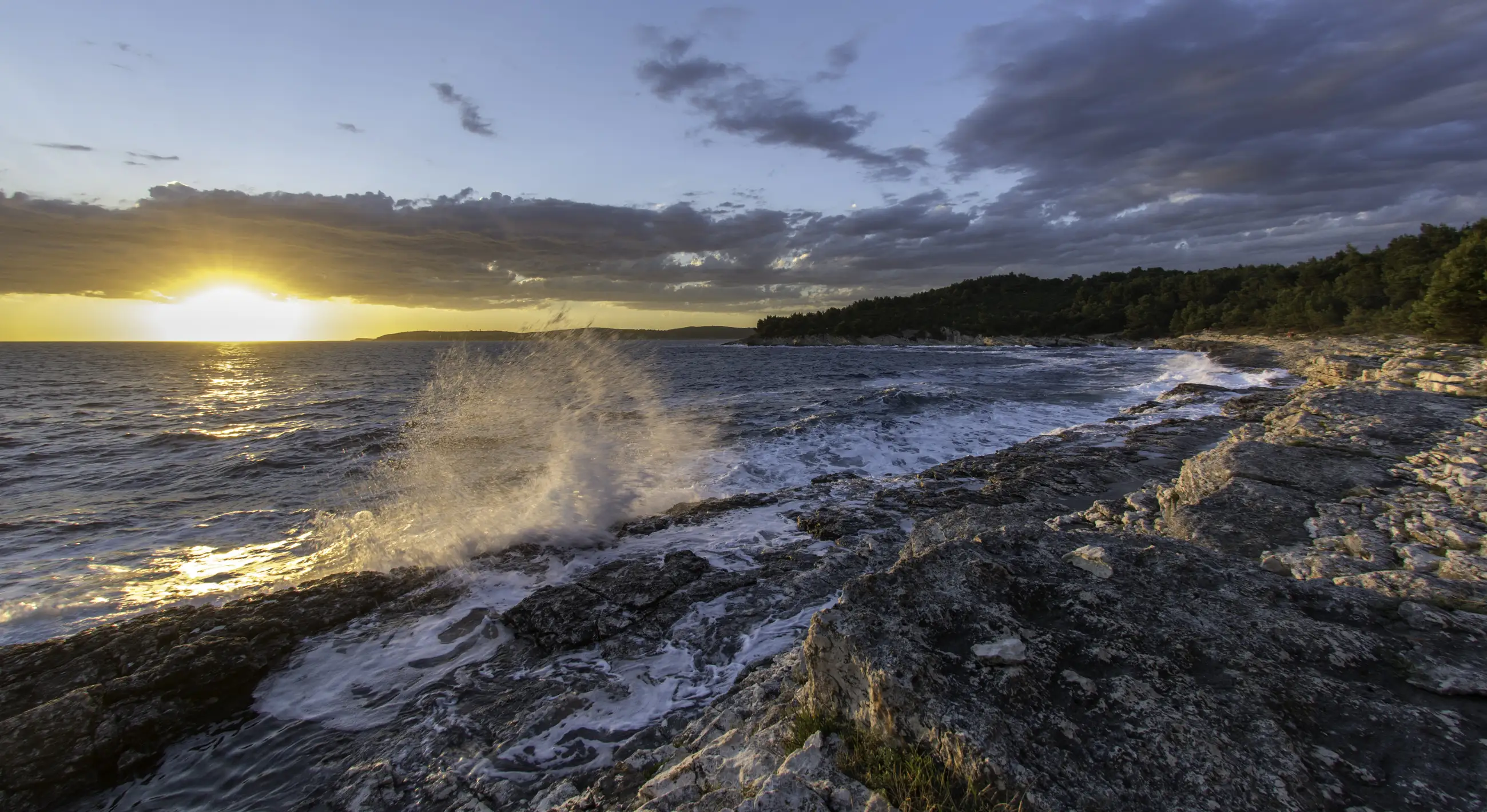 Waves crash against rocky shore at sunset, sending spray into the air. The sun is low on the horizon, casting warm golden light on the ocean and clouds, with rocks and tide pools in the foreground.