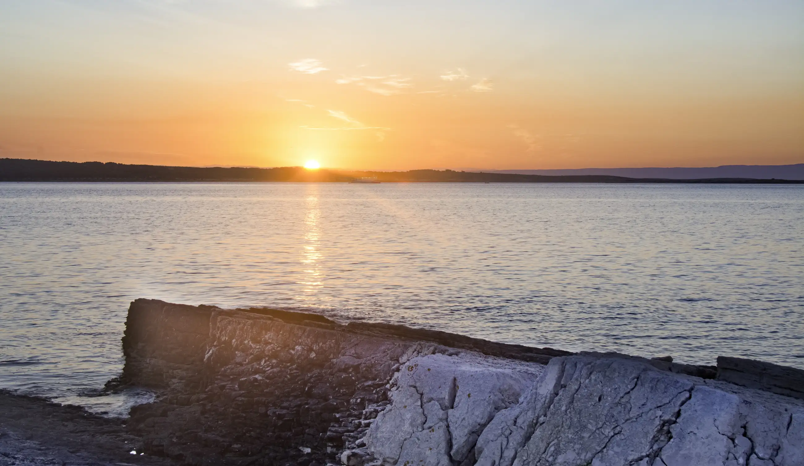 A serene sunset over a calm body of water, with the sun low on the horizon, casting orange and pink hues across the sky and reflecting on the water. Rocky shorelines are visible in the foreground.