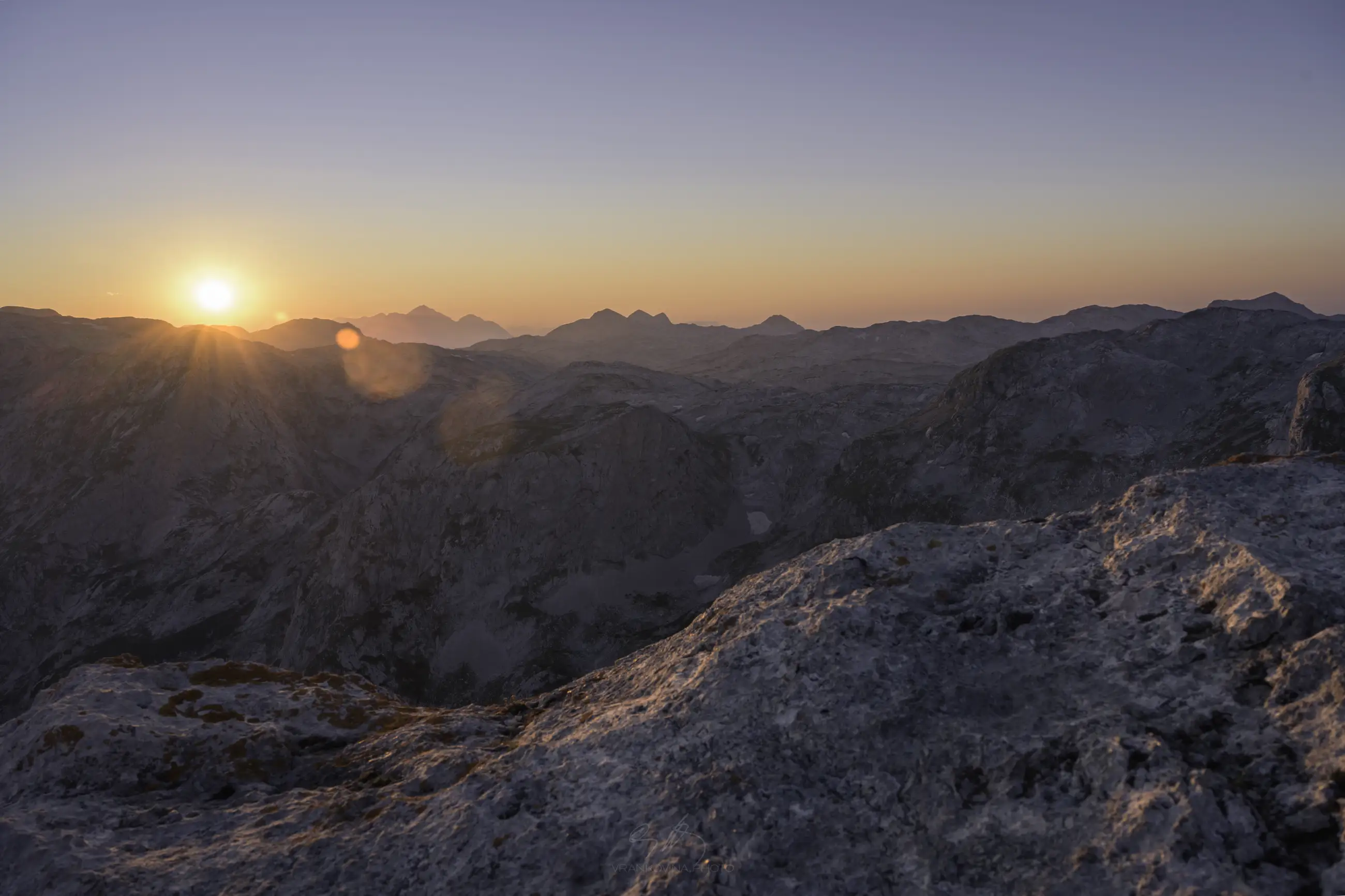Sun rising over distant mountain peaks, casting a warm orange glow across the rocky landscape and illuminating the sky with soft pastel colors. Foreground features rugged rocks with visible texture.
