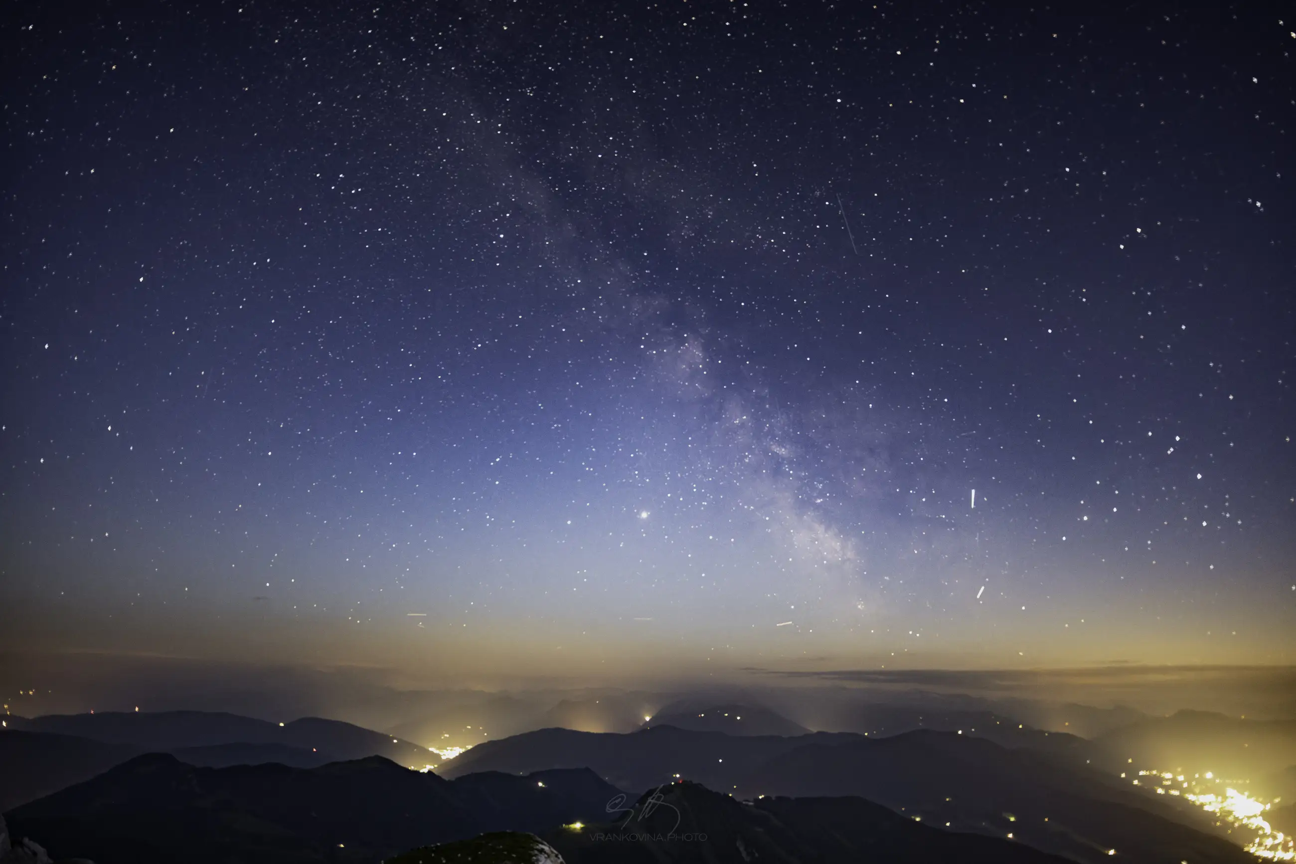 A star-filled night sky and the Milky Way stretch over silhouetted mountains, with faint lights glowing in valleys below and a subtle gradient of blue and orange near the horizon.