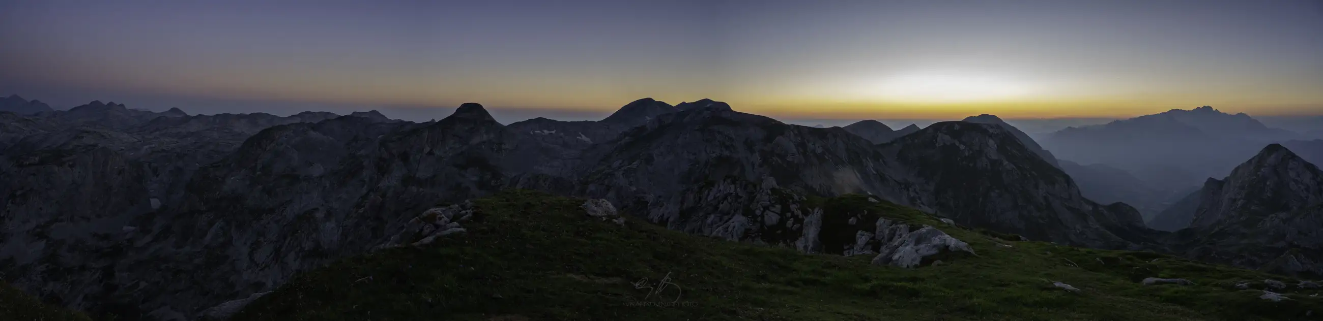 A panoramic view of rugged mountain peaks under a clear sky at sunrise, with a soft orange glow on the horizon and rocky terrain in the foreground.