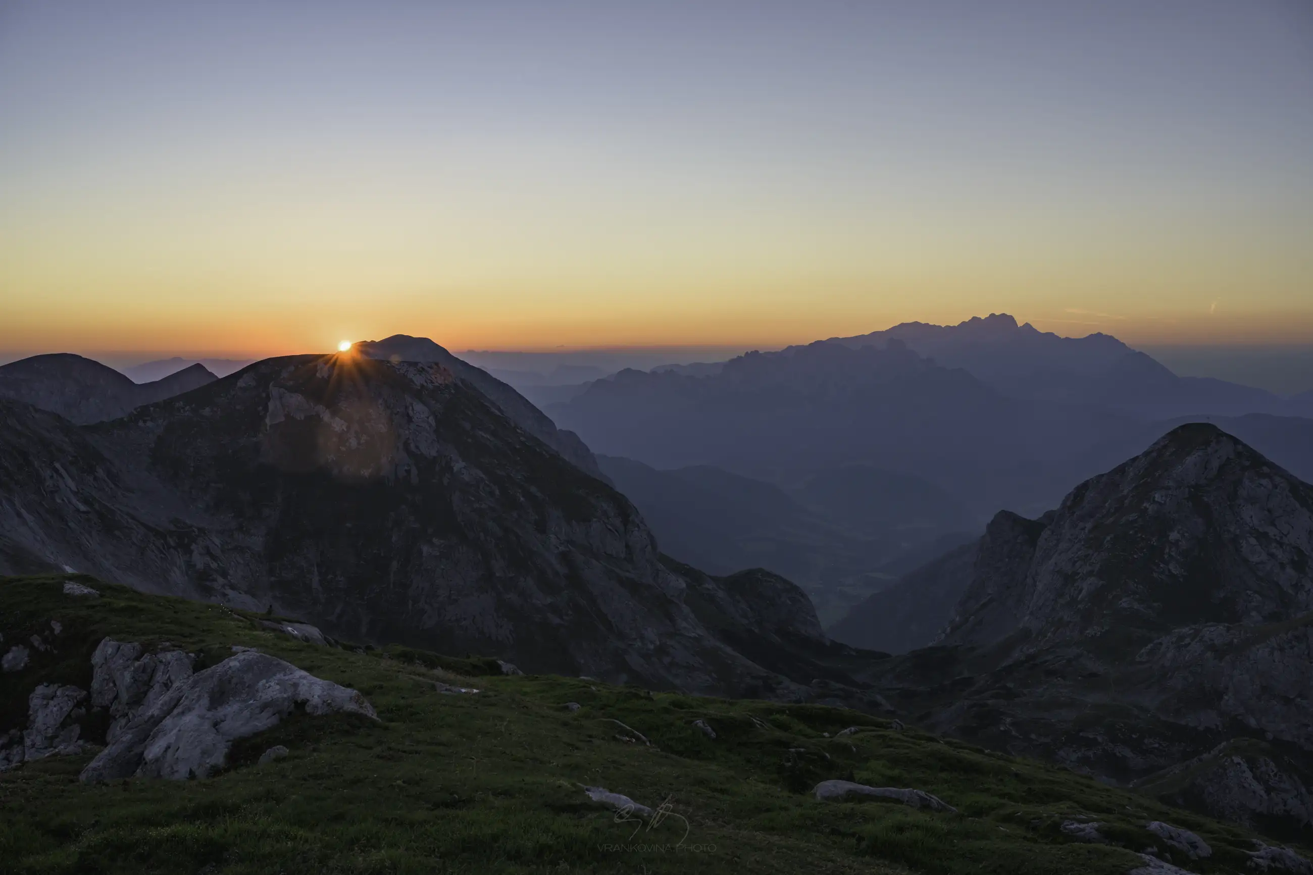 The sun rises behind a mountain peak, casting warm light over grassy hills and rocky slopes, with misty blue mountains fading into the distance under a clear sky.