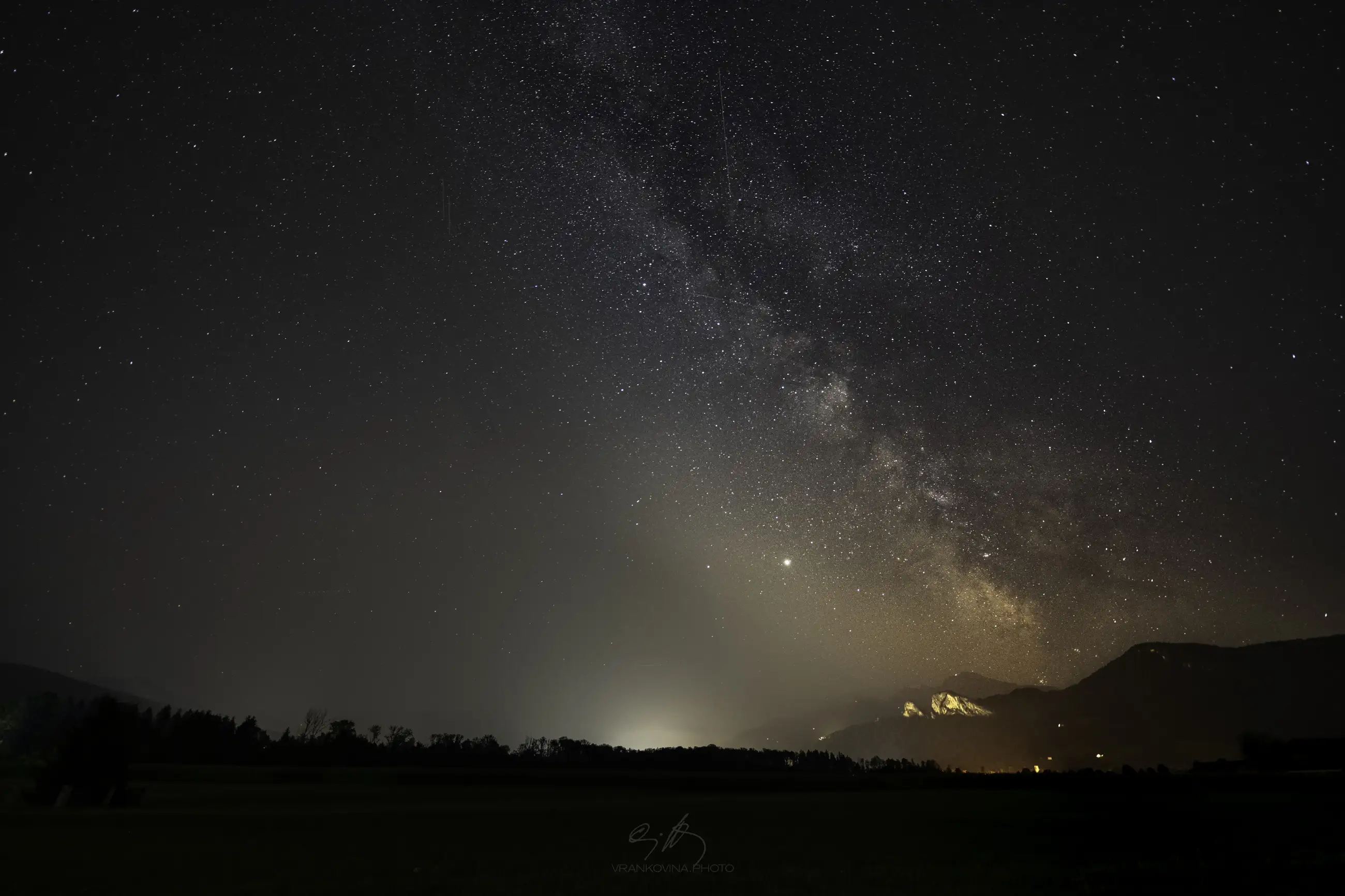 A clear night sky filled with stars and the glowing band of the Milky Way is visible above a dark landscape with distant mountains and faint lights near the horizon.