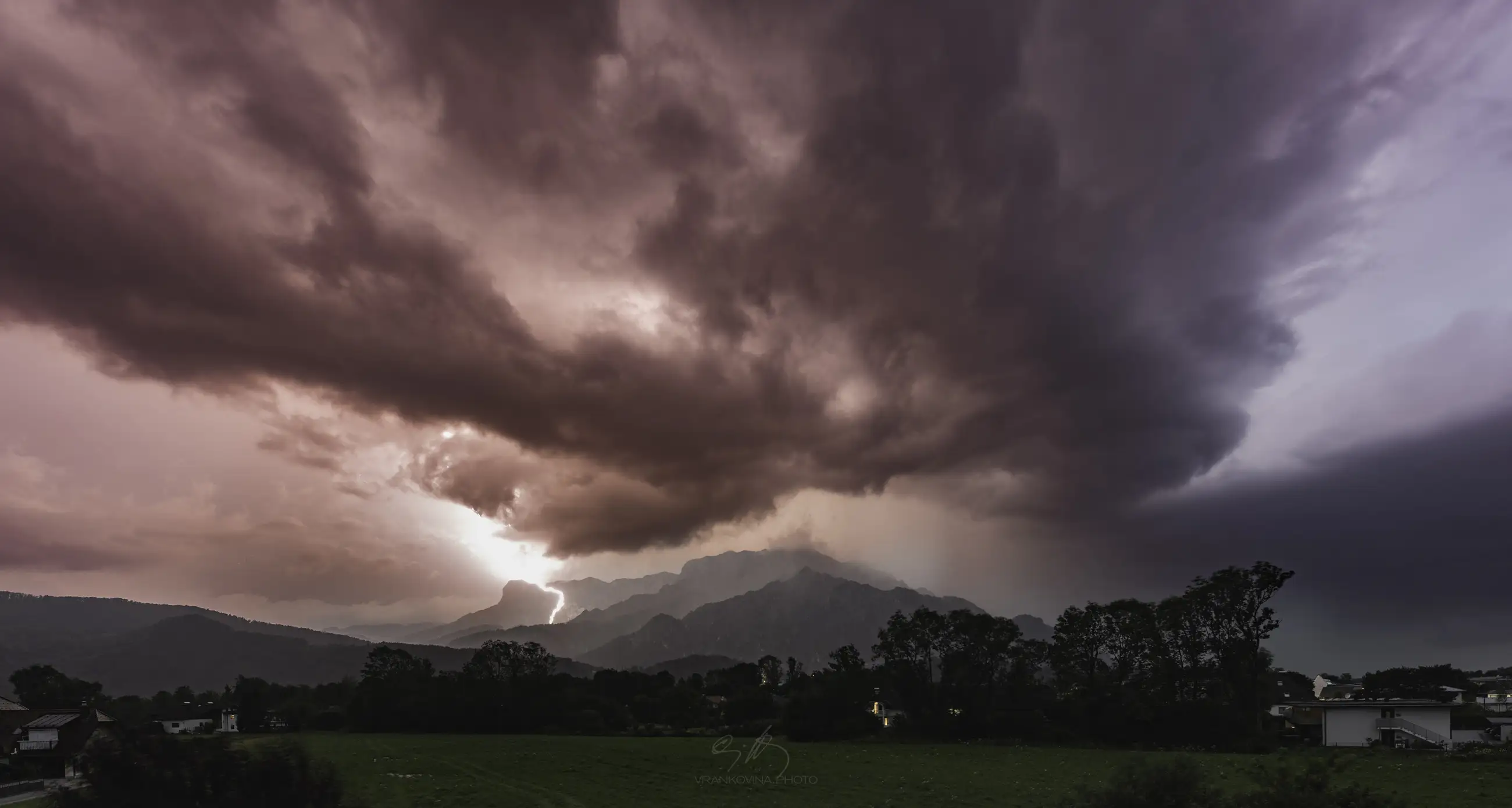 A dramatic sky shows lightning striking behind silhouetted trees and buildings, with dark storm clouds on the left and an orange glow illuminating the scene. Mountains are faintly visible in the background.