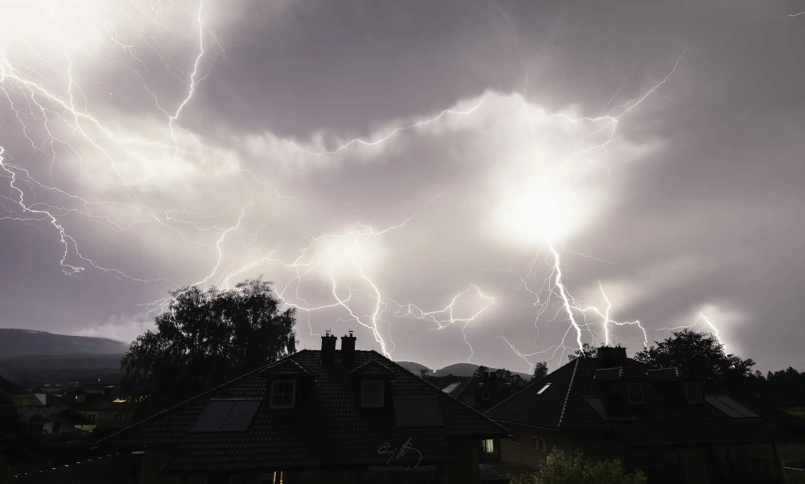 Bright lightning bolts streak across a cloudy night sky above dark silhouetted houses and trees in a residential area, illuminating the entire scene dramatically.