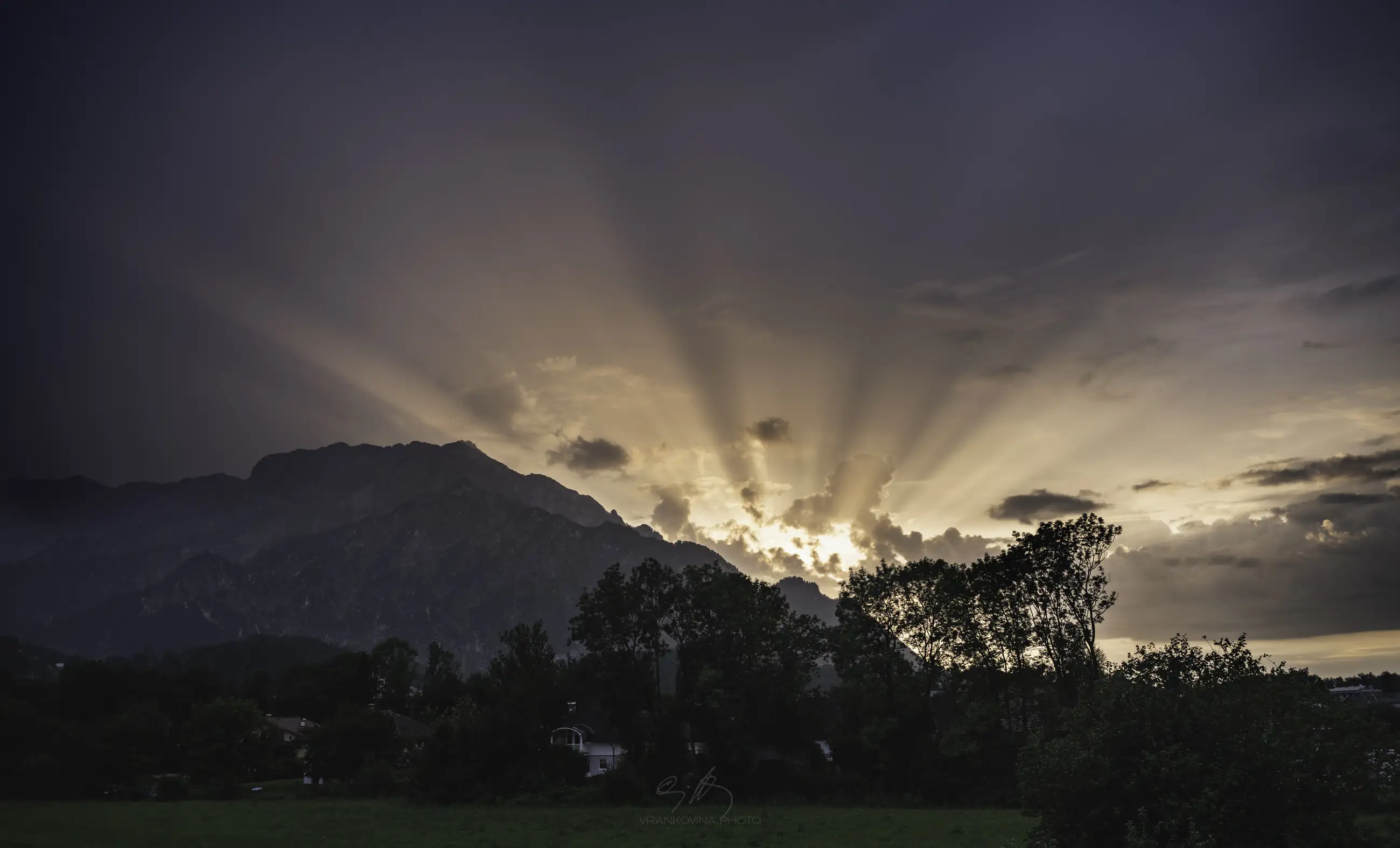 Sunset behind a mountain range with dramatic sun rays streaming through clouds, silhouetting trees and casting a soft glow over a green landscape in the foreground.